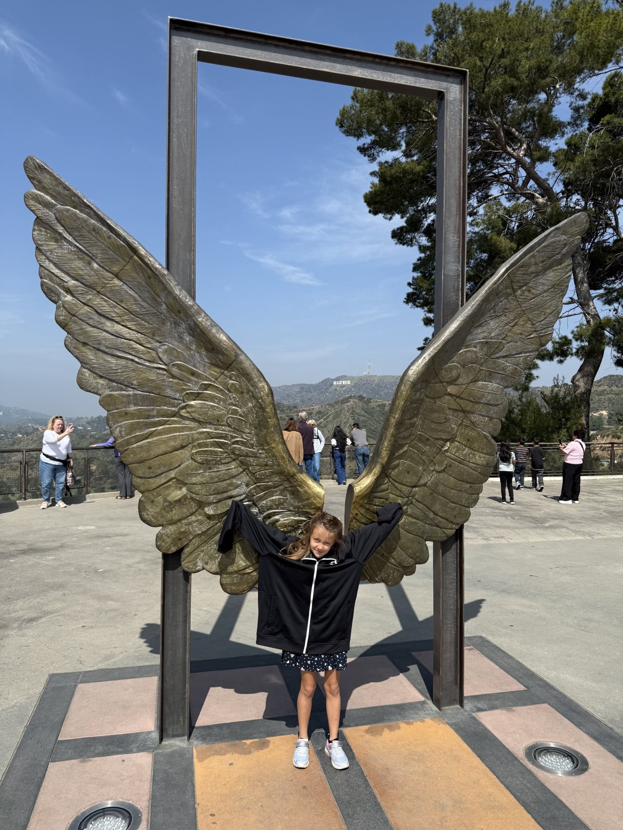 Dívka stojí mezi bronzovými křídly s výhledem na Hollywood Sign.