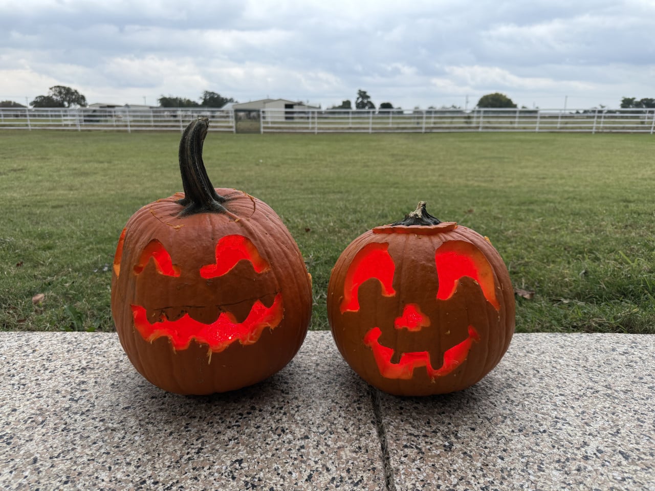 Dvě rozsvícené halloweenské dýně na kamenné terase před loukou a plotem