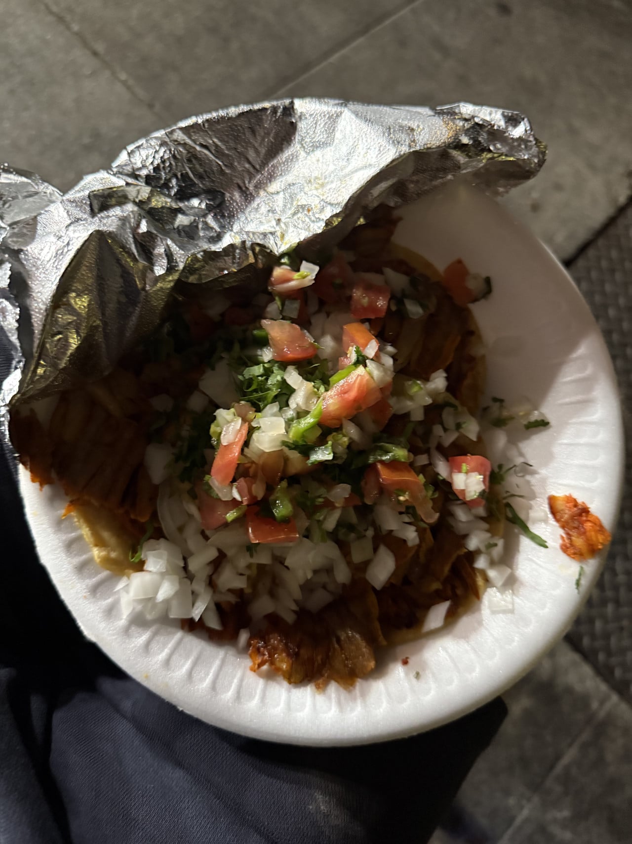 Papírová miska s tacos pod alobalem, posypaná cibulí, rajčaty a koriandrem.