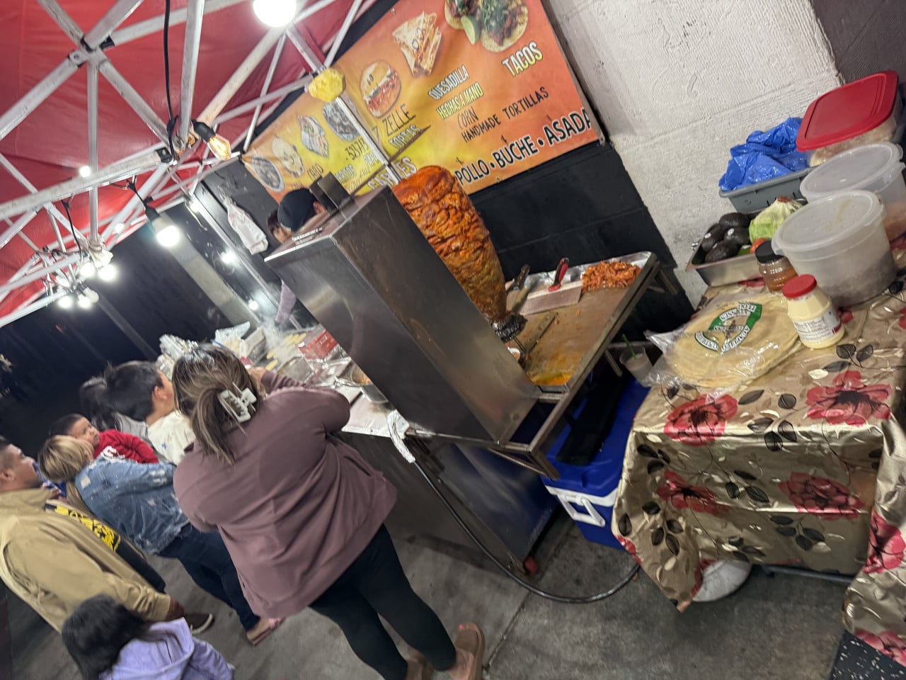 Pouliční stánek s tacos, vertikální gril a fronta lidí pod červeným stanem.