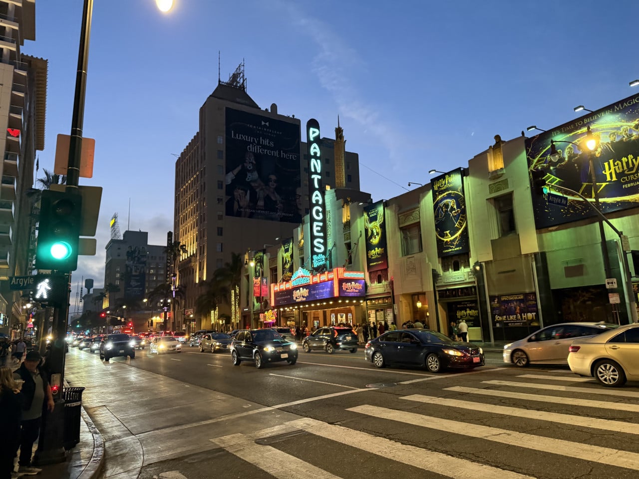 Večerní ulice Hollywoodu s neony, auty a nápisem Pantages Theatre.