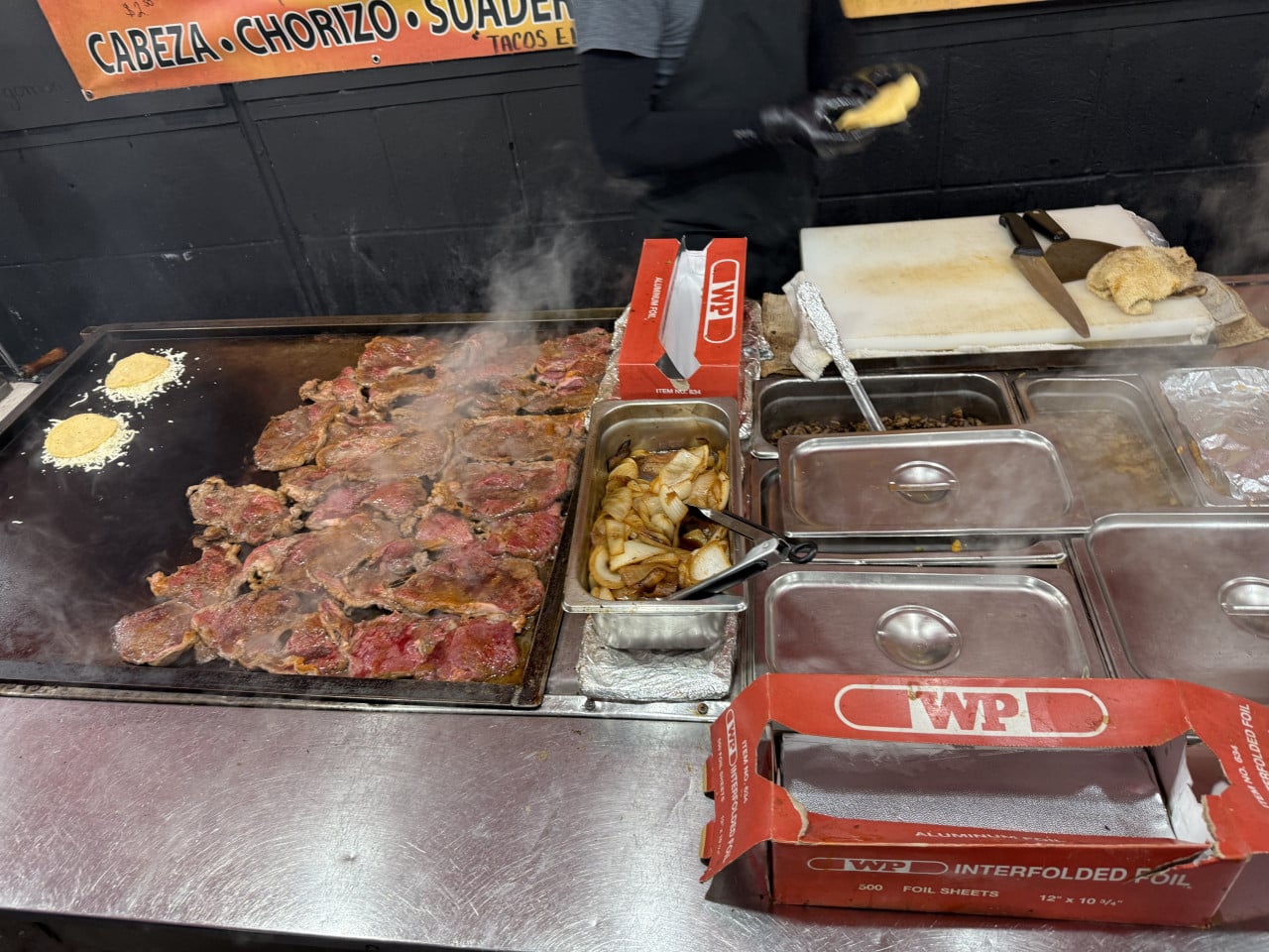 Na grilu se opéká maso, cibule a tortilly vedle kuchařského pultu.