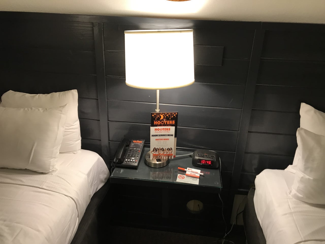 Noční stolek mezi postelemi, lampa, telefon, budík 10:46 a HOOTERS ROOM SERVICE MENU.