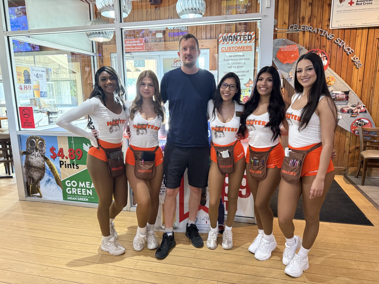 Muž pózuje s pěti servírkami Hooters; za nimi nápisy WANTED CUSTOMERS, $4.89.