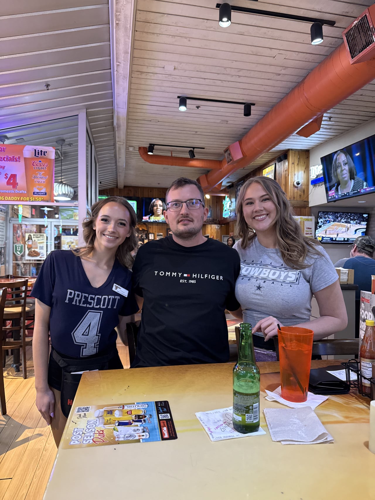 Muž u stolu s lahví piva, vedle servírky PRESCOTT 4 a DALLAS COWBOYS.