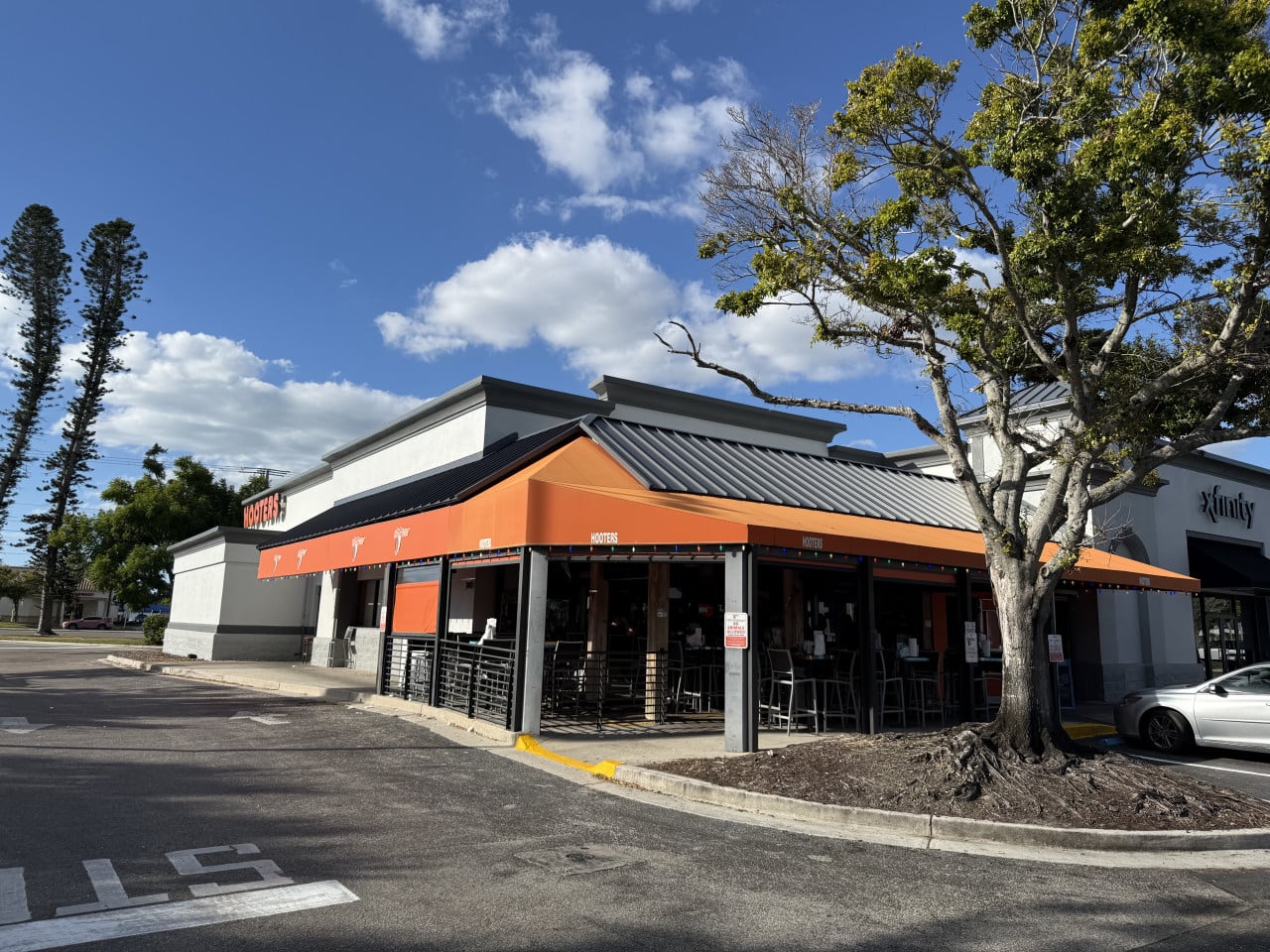 Exteriér restaurace HOOTERS s oranžovými markýzami, parkovištěm, stromem a nápisem Xfinity.