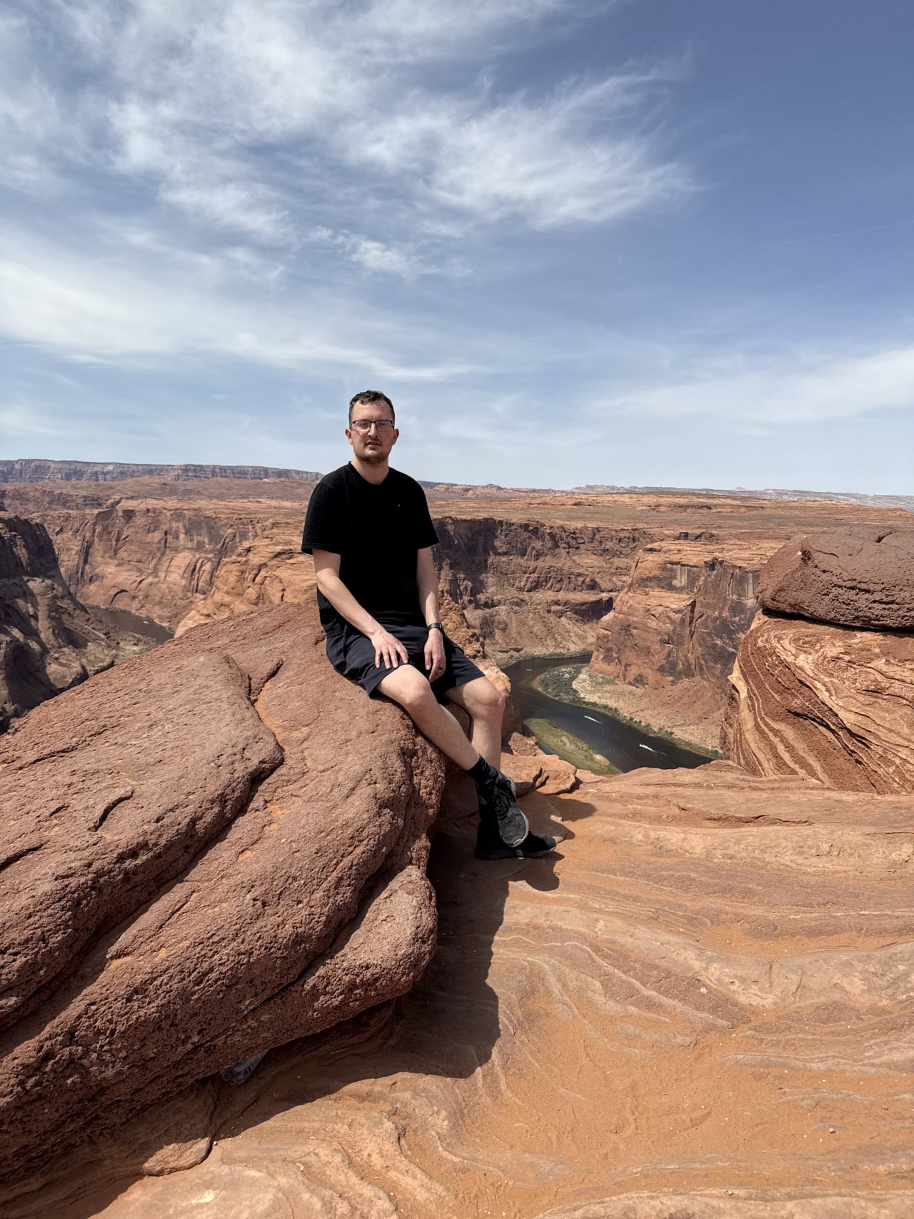 Muž sedí na skalní římse, za ním kaňon a ohyb řeky Colorado.