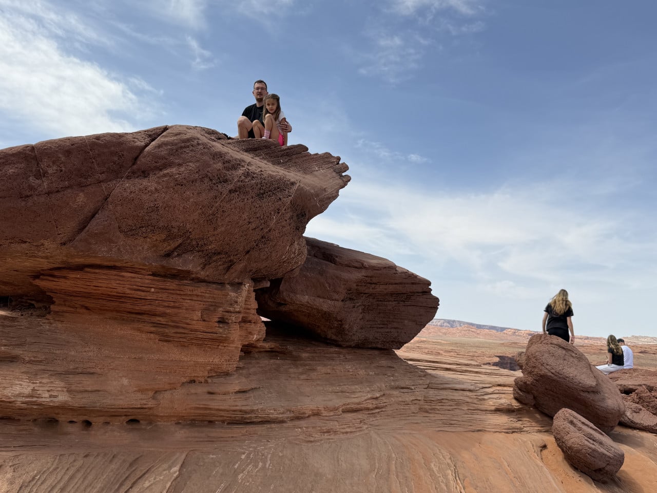 Muž s dívkou sedí na skalním převisu, vpravo turisté v poušti pod modrou oblohou