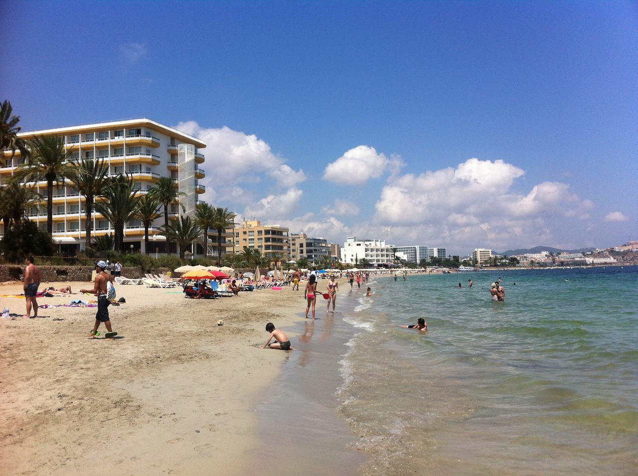 Pláž s lidmi, slunečníky a hotely u moře, modrá obloha s mraky