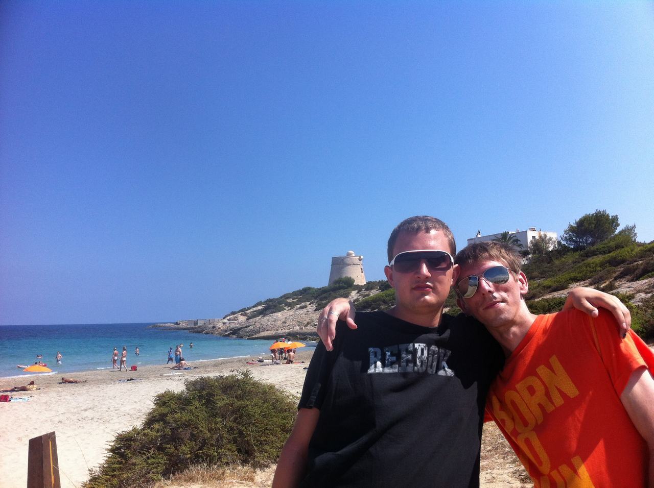 Dva muži v brýlích na pláži, trička REEBOK a BORN, moře v pozadí.