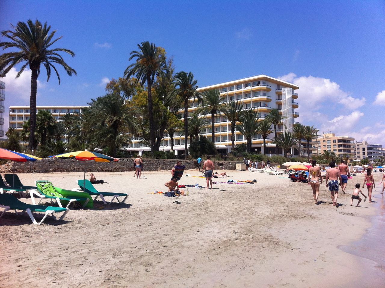 Písečná pláž s lehátky a slunečníky, lidé u moře, palmy a hotel.