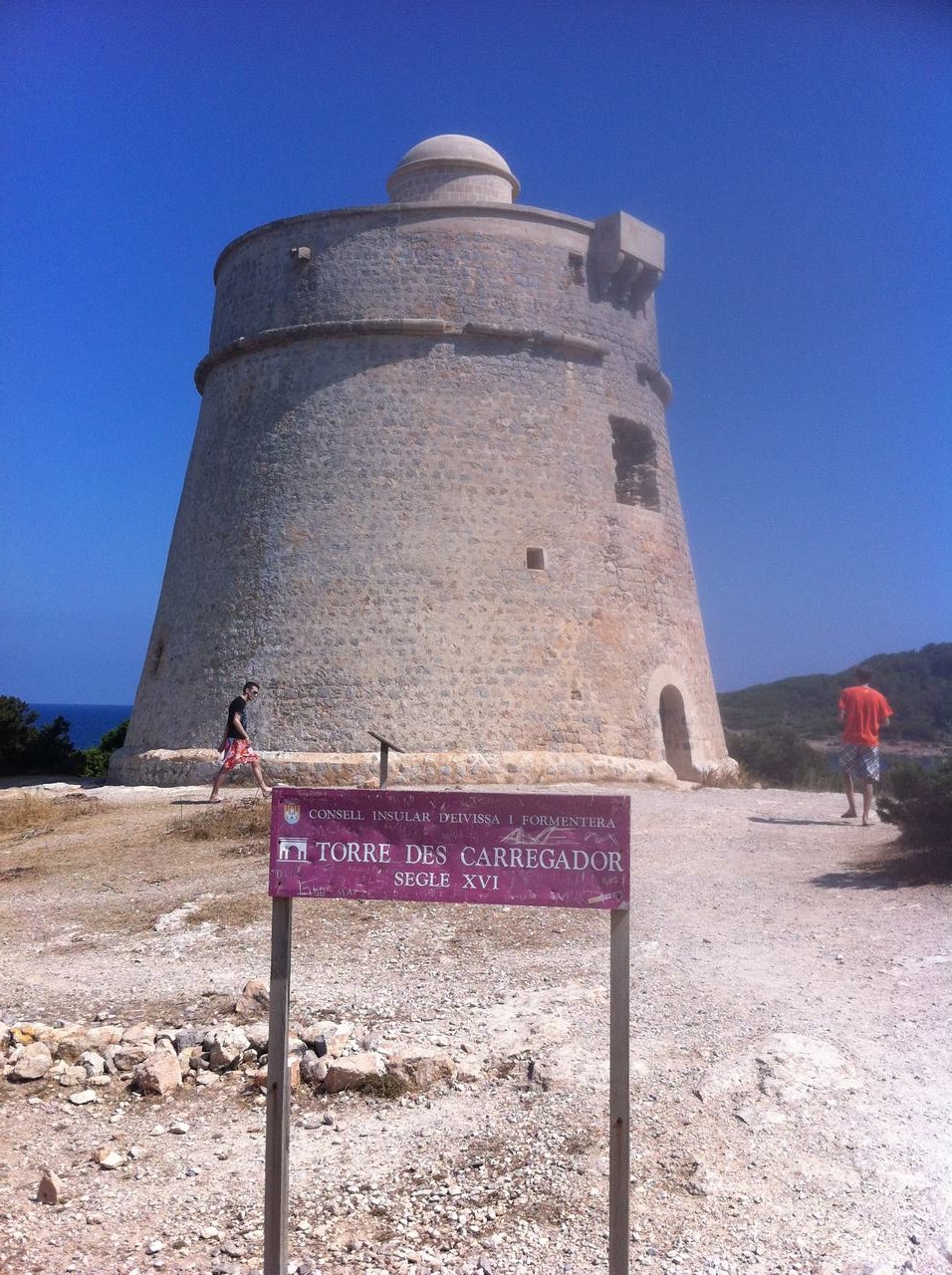 Kamenná věž a cedule TORRE DES CARREGADOR SEGLE XVI, dva turisté