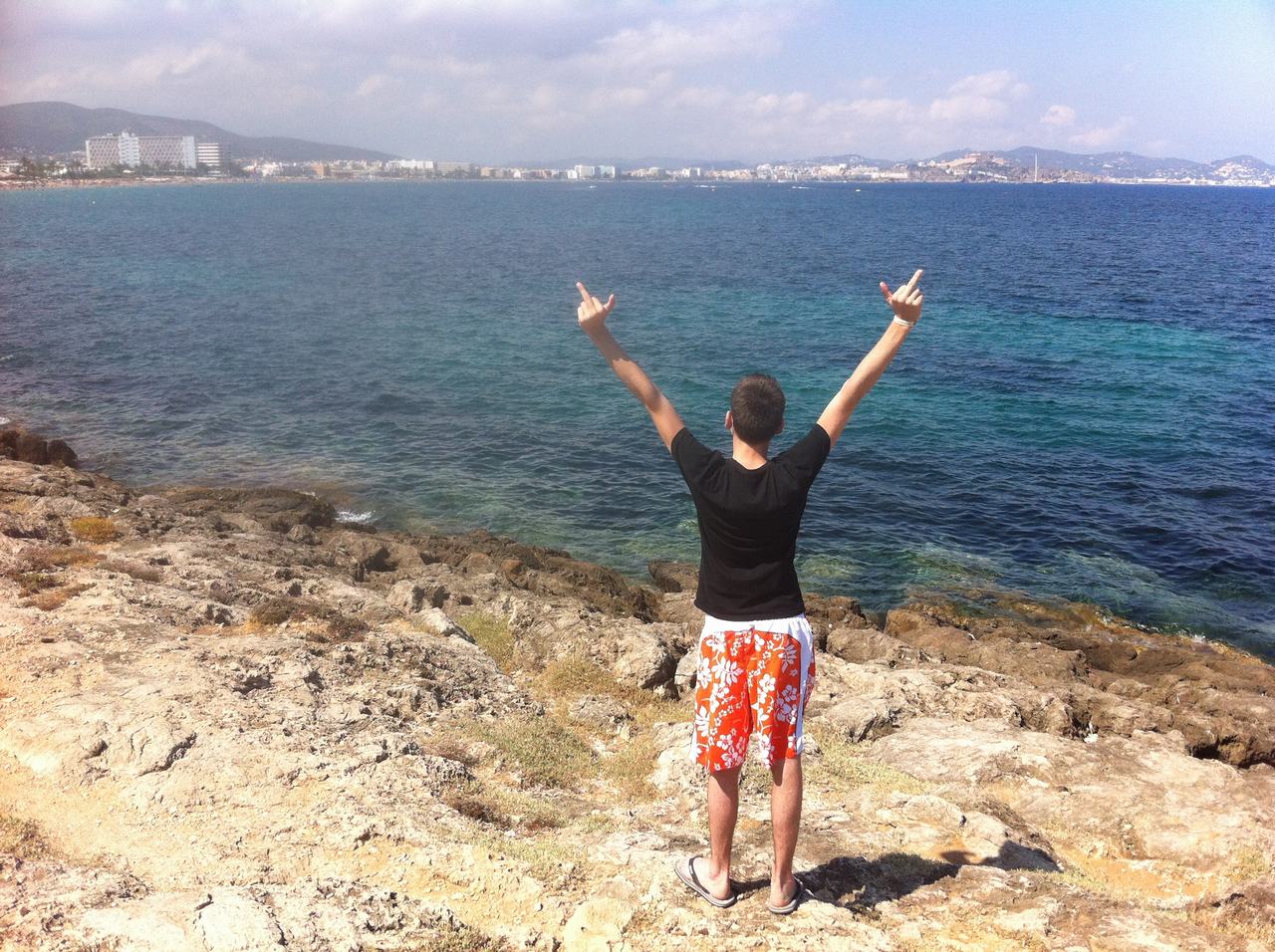 Muž na skalnatém pobřeží s oběma zdviženými prostředníčky hledí na moře