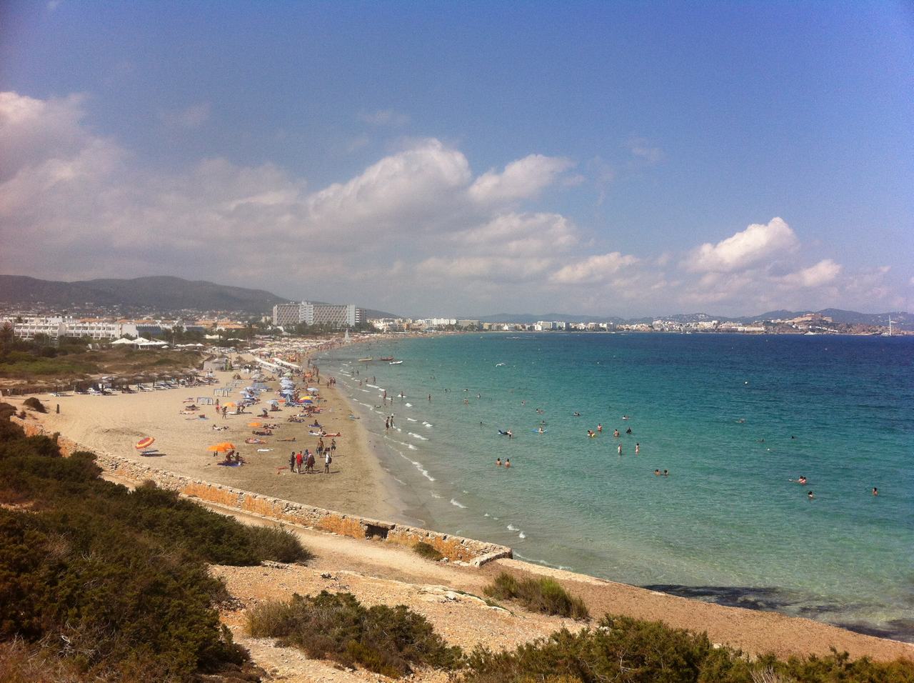 Panoramatický pohled na pláž s koupajícími se lidmi, tyrkysovým mořem a hotely v dálce