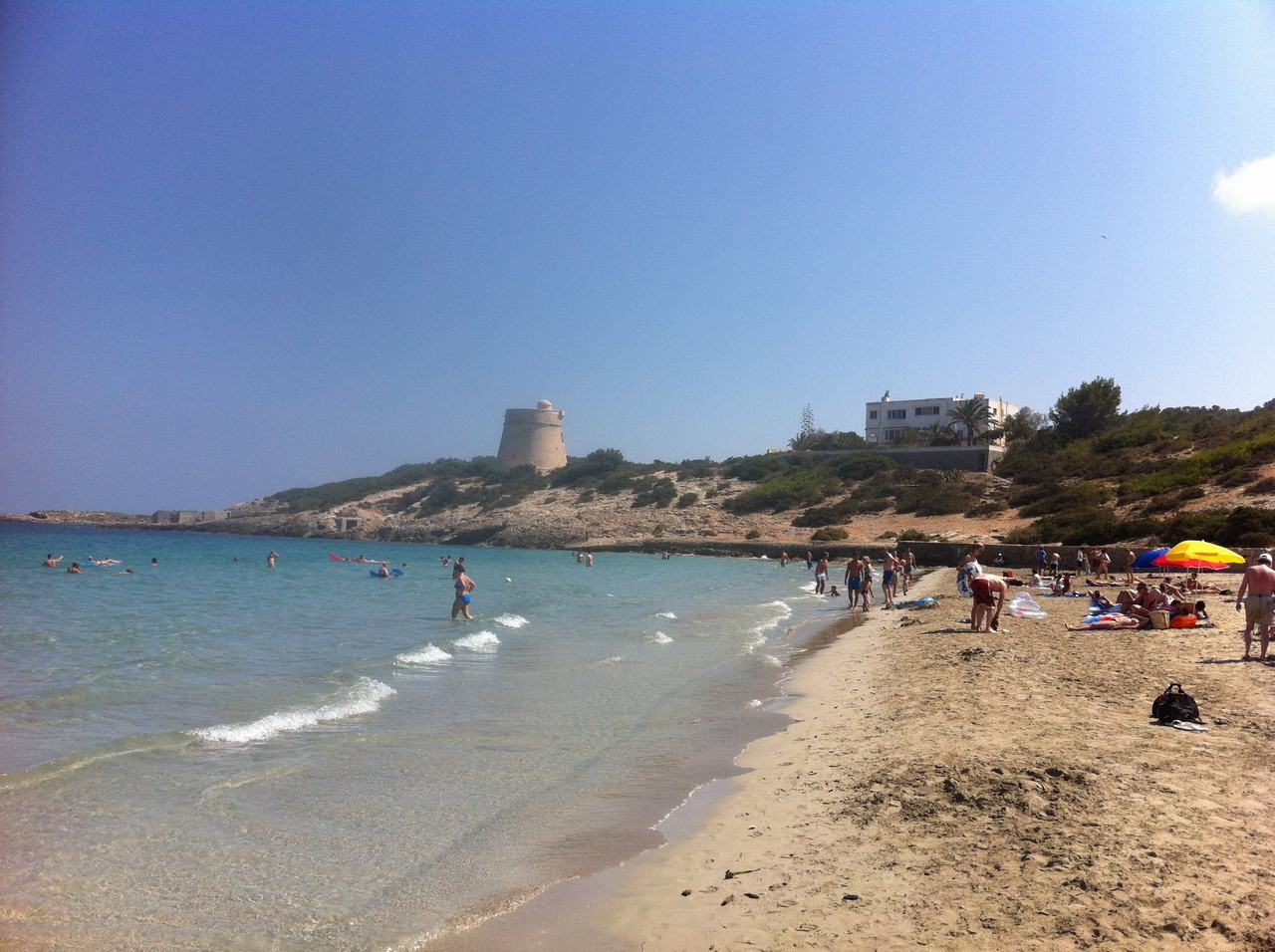 Písečná pláž s koupajícími se lidmi, tyrkysové moře, věž a bílé domy v dálce