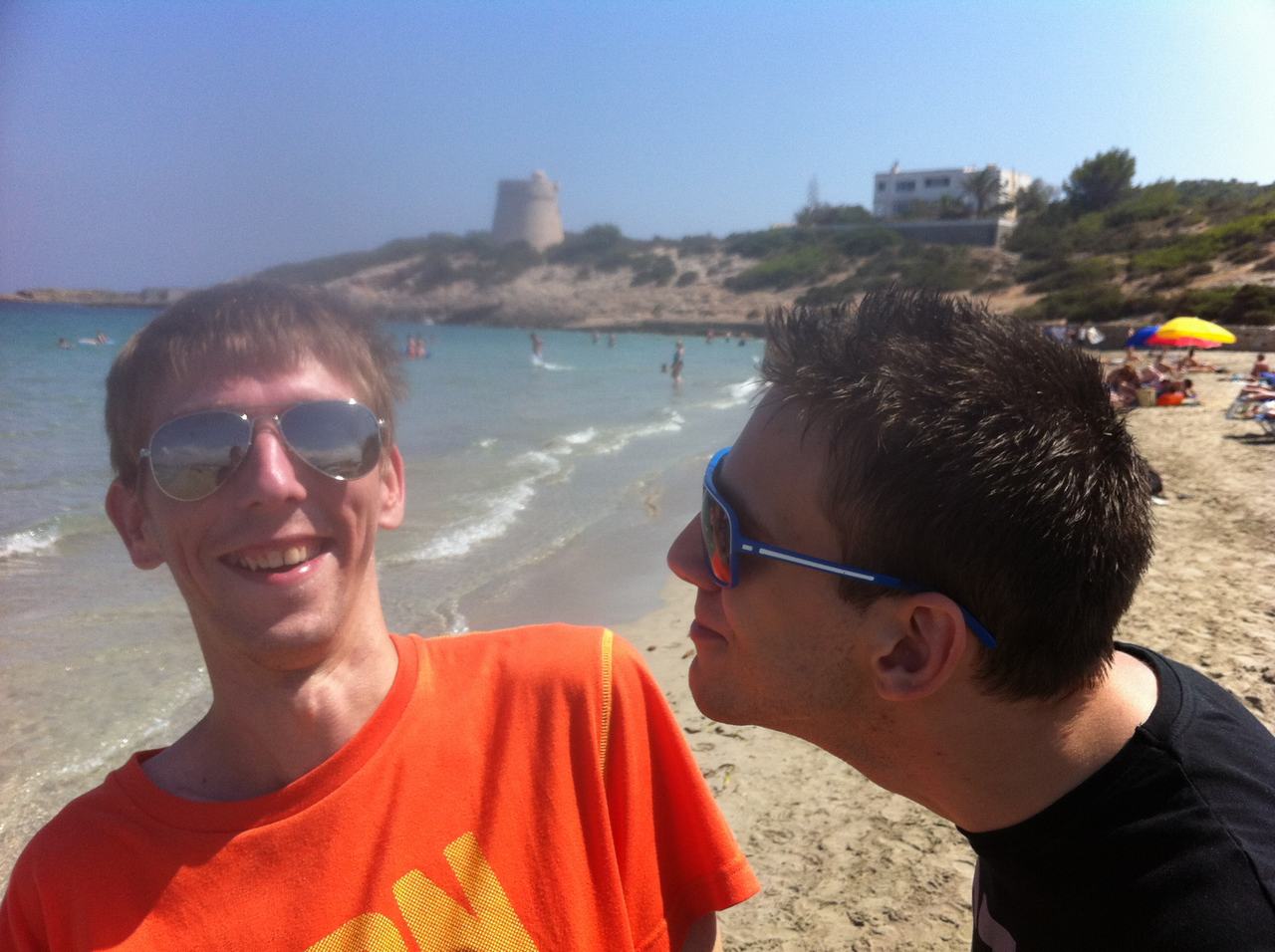 Dva kamarádi v brýlích se fotí na pláži, v pozadí moře a plavci