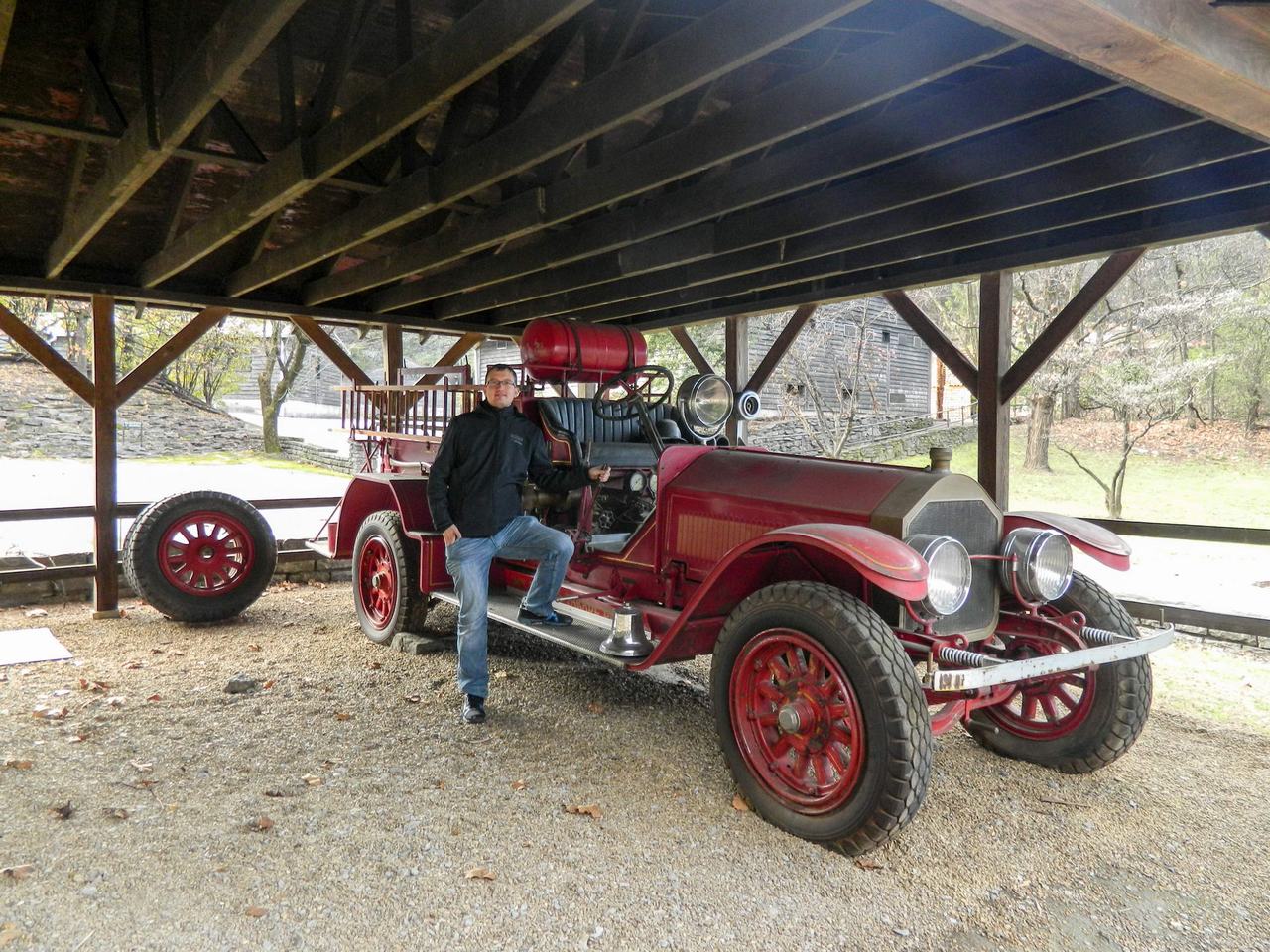 Muž pózuje u historického červeného hasičského vozu pod dřevěným přístřeškem.