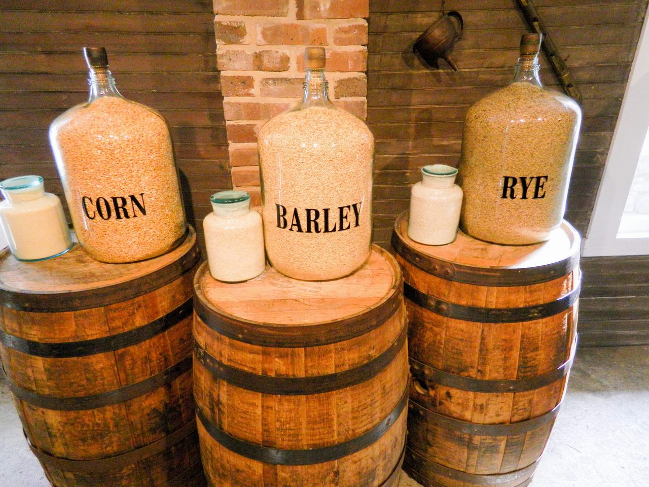Skleněné demižony s nápisy CORN, BARLEY a RYE na dřevěných sudech.