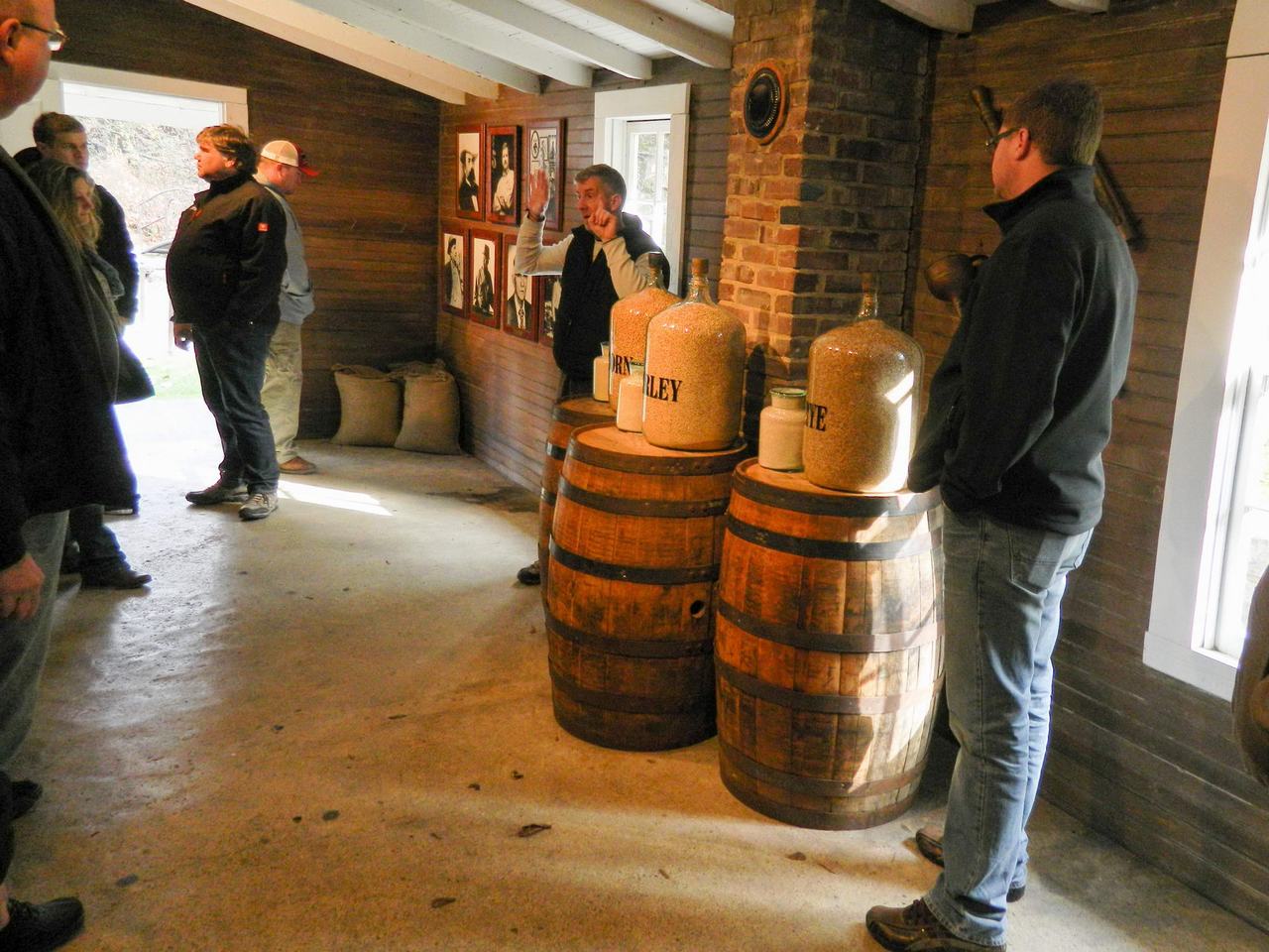 Průvodce mluví ke skupině v dřevěné místnosti; sudy s džbány FARM VALLEY.