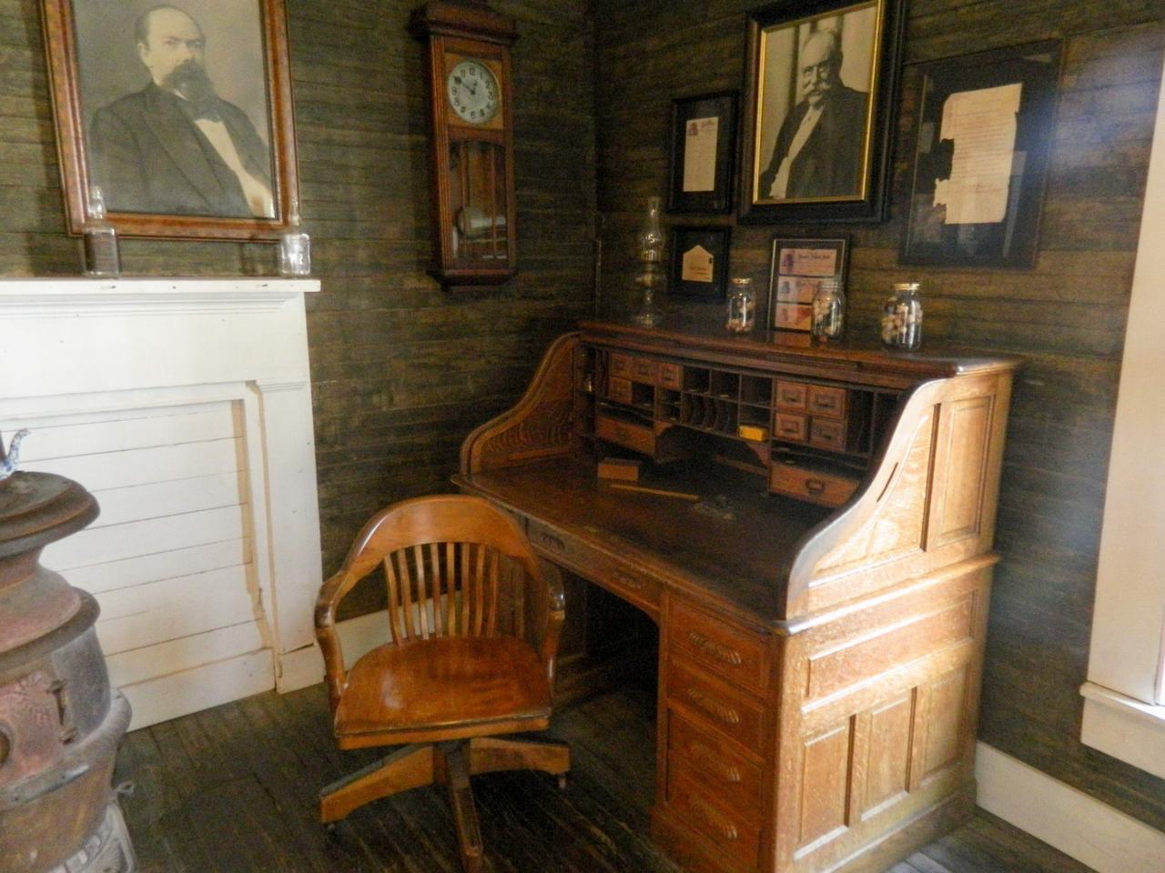 Starý dřevěný psací stůl se židlí, krb, nástěnné portréty a hodiny.