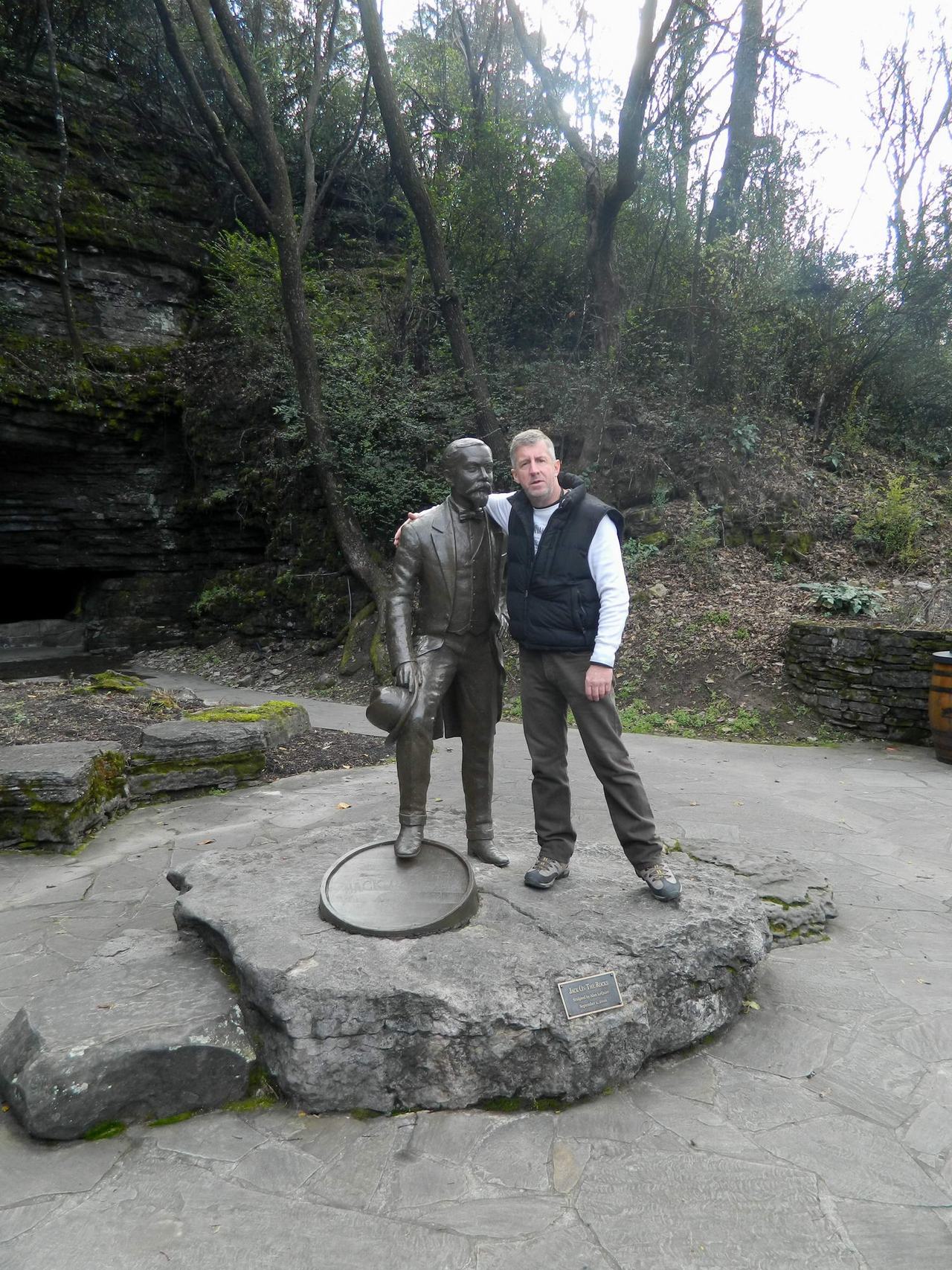 Muž objímá bronzovou sochu muže na kamenném podstavci u skály v lese.