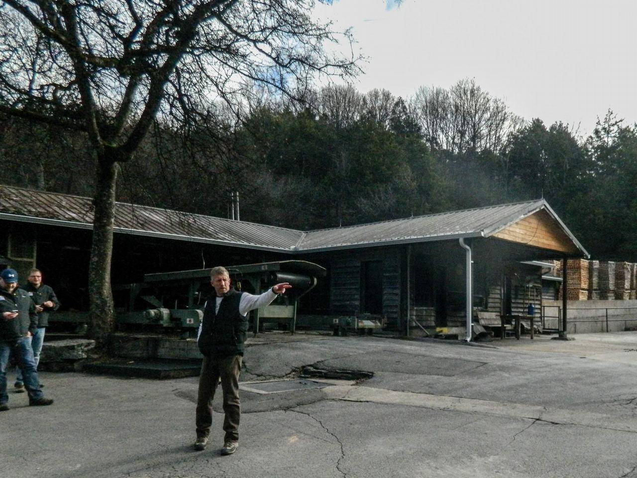 Průvodce ukazuje směr před dřevěnými budovami destilerie, v pozadí les