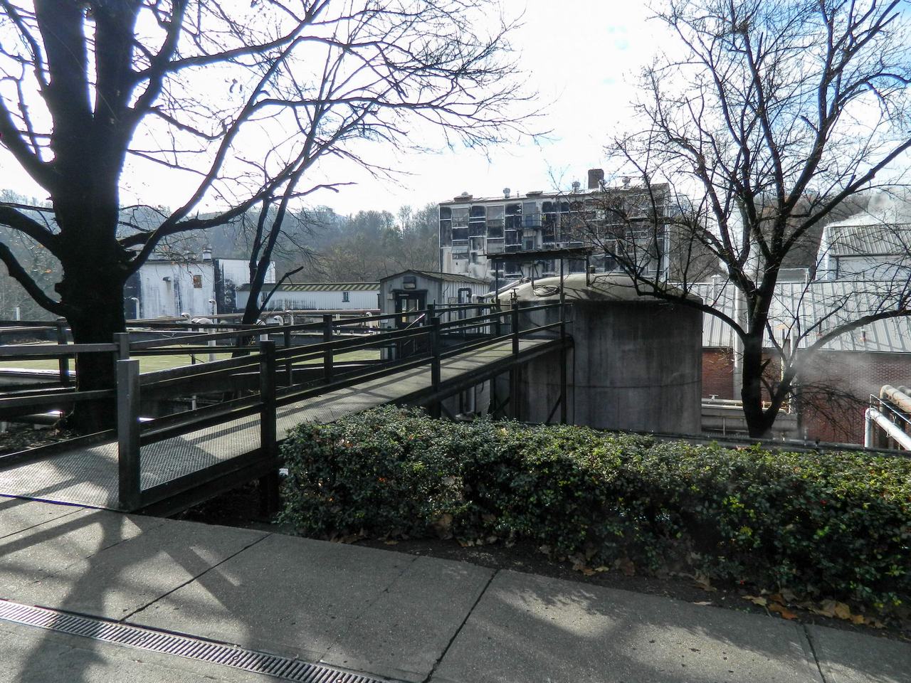 Mostek se zábradlím vede k budovám destilerie, v pozadí stoupá pára