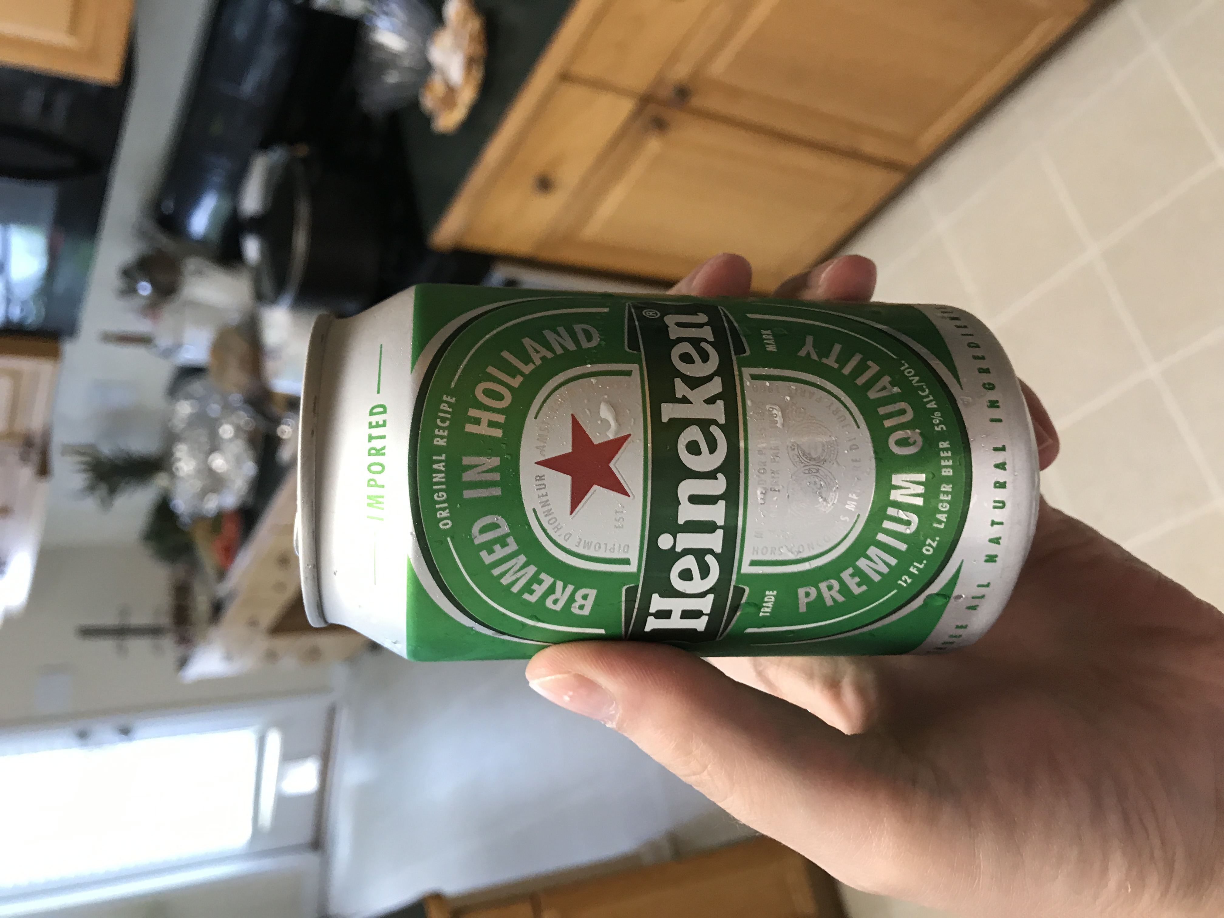 Ruka drží plechovku piva Heineken, Brewed in Holland, Premium Quality, v kuchyni.