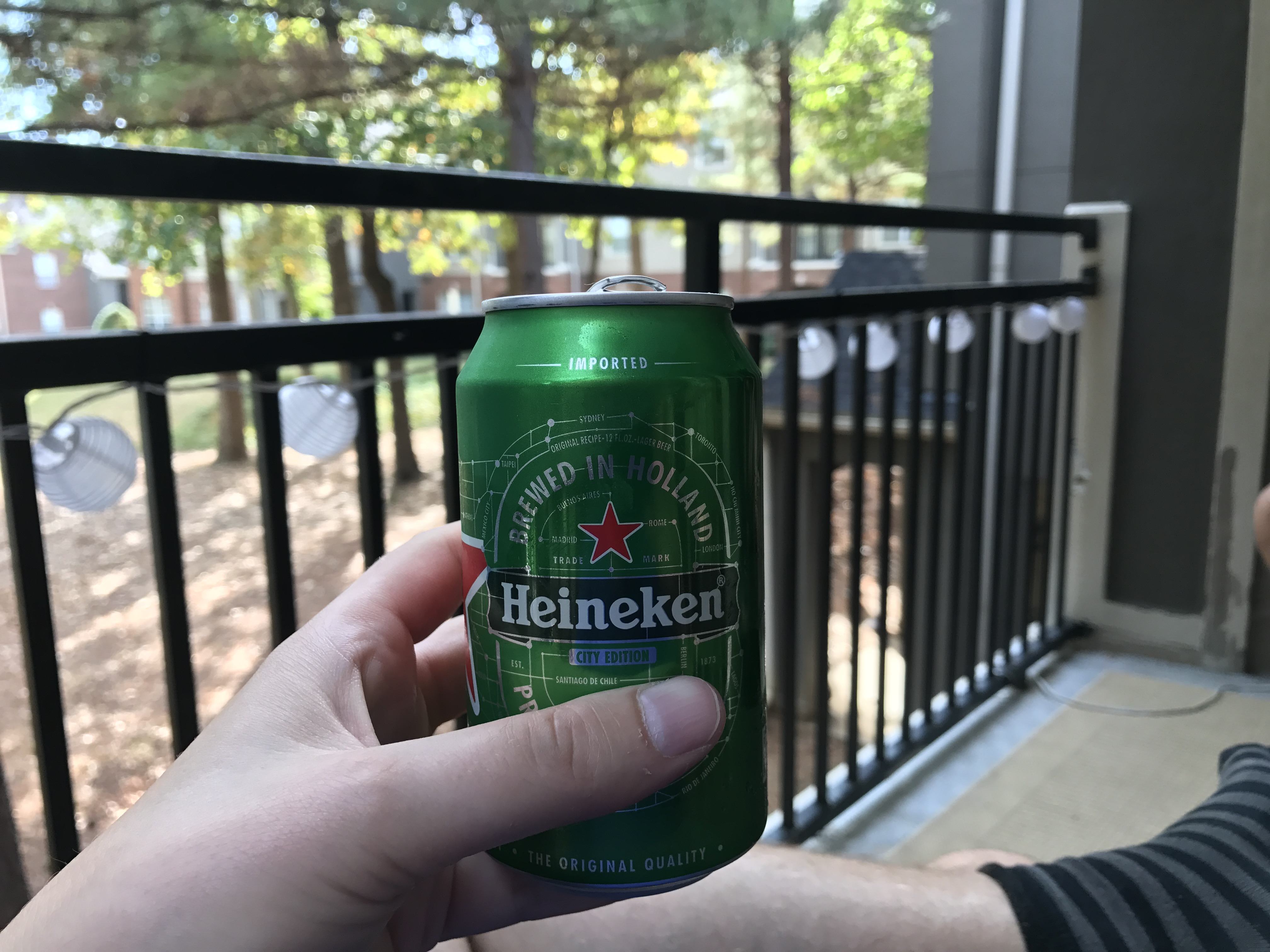 Ruka drží plechovku Heineken, nápisy Imported a City Edition, balkón v pozadí.