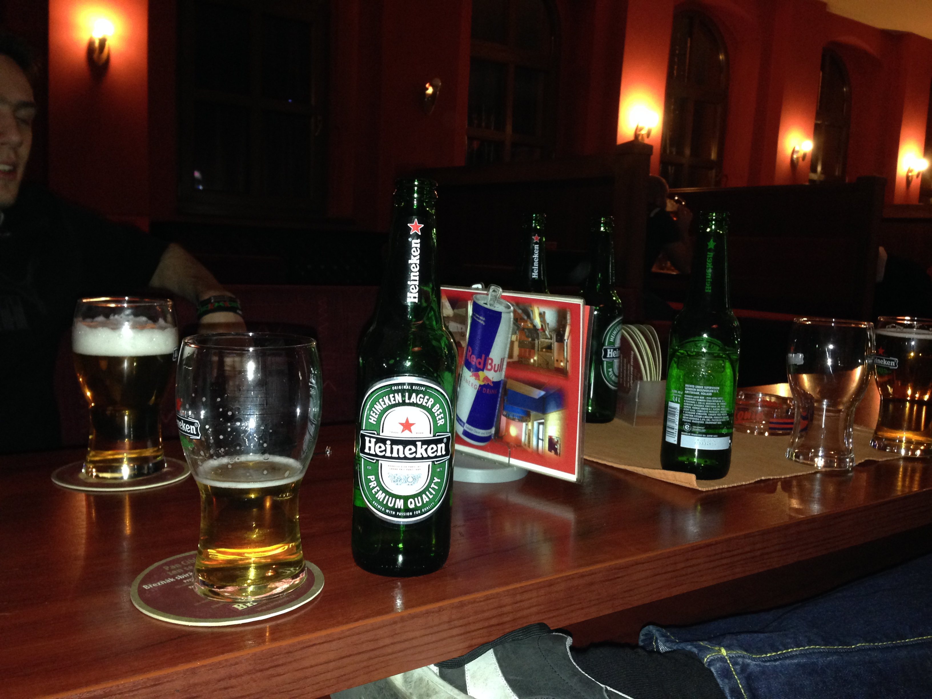Stůl v hospodě s lahvemi Heineken, sklenicemi piva a plechovkou Red Bull.