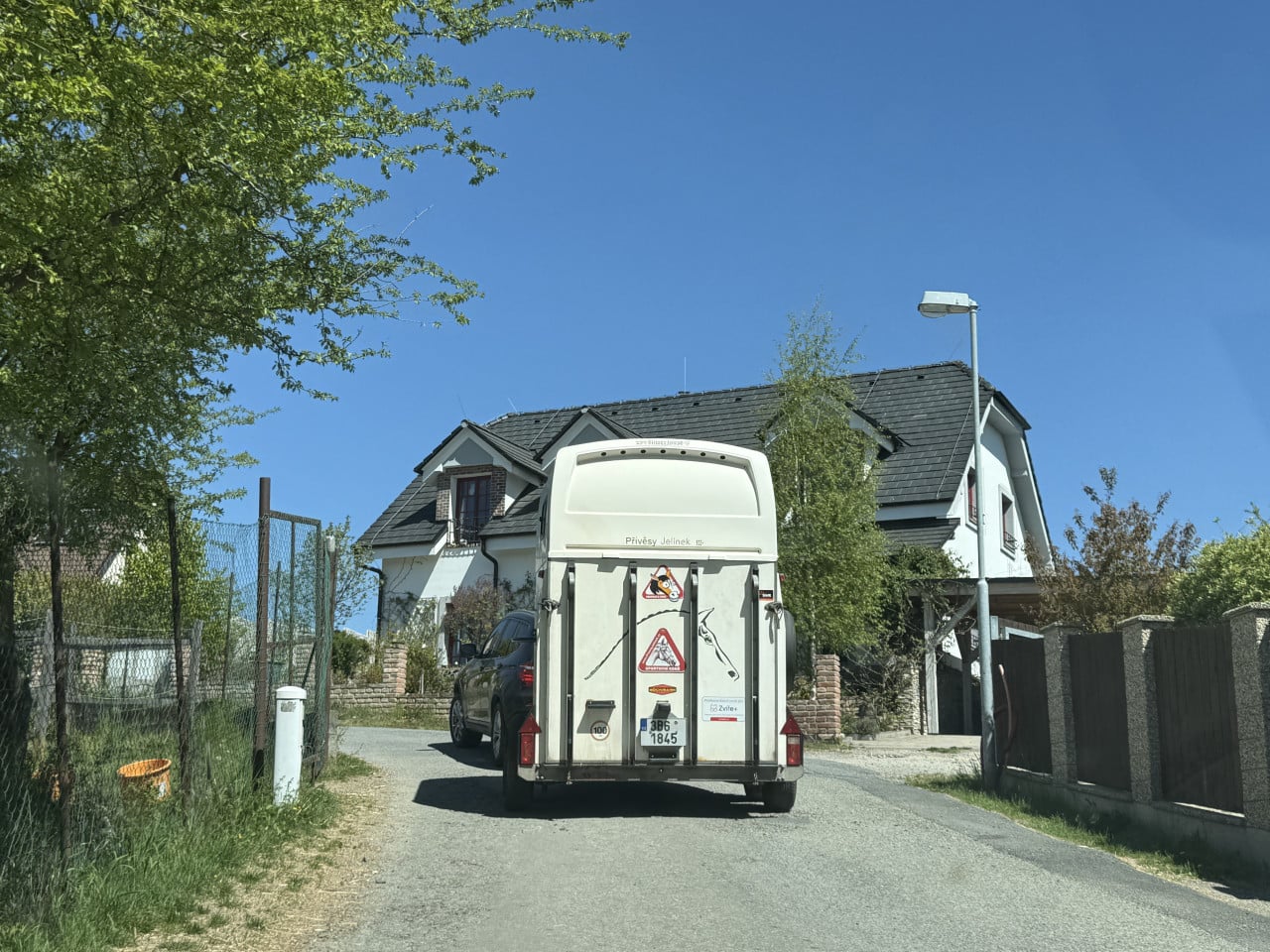 Přívěs s koňmi u domu na úzké silnici, modrá obloha a stromy.