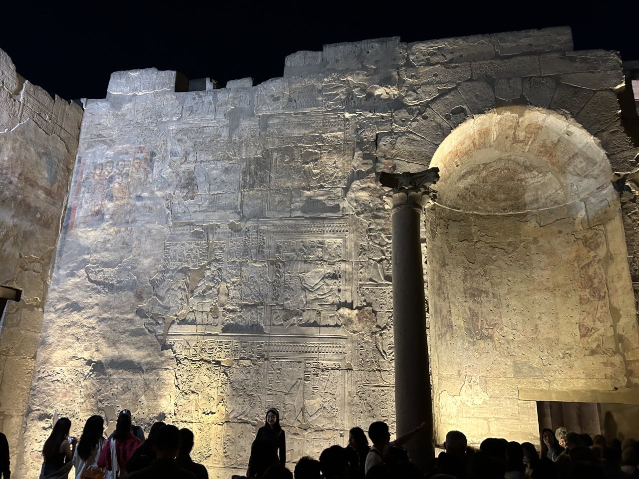 Osvětlená reliéfní stěna chrámu Karnak s obloukem a sloupem, dole turisté.