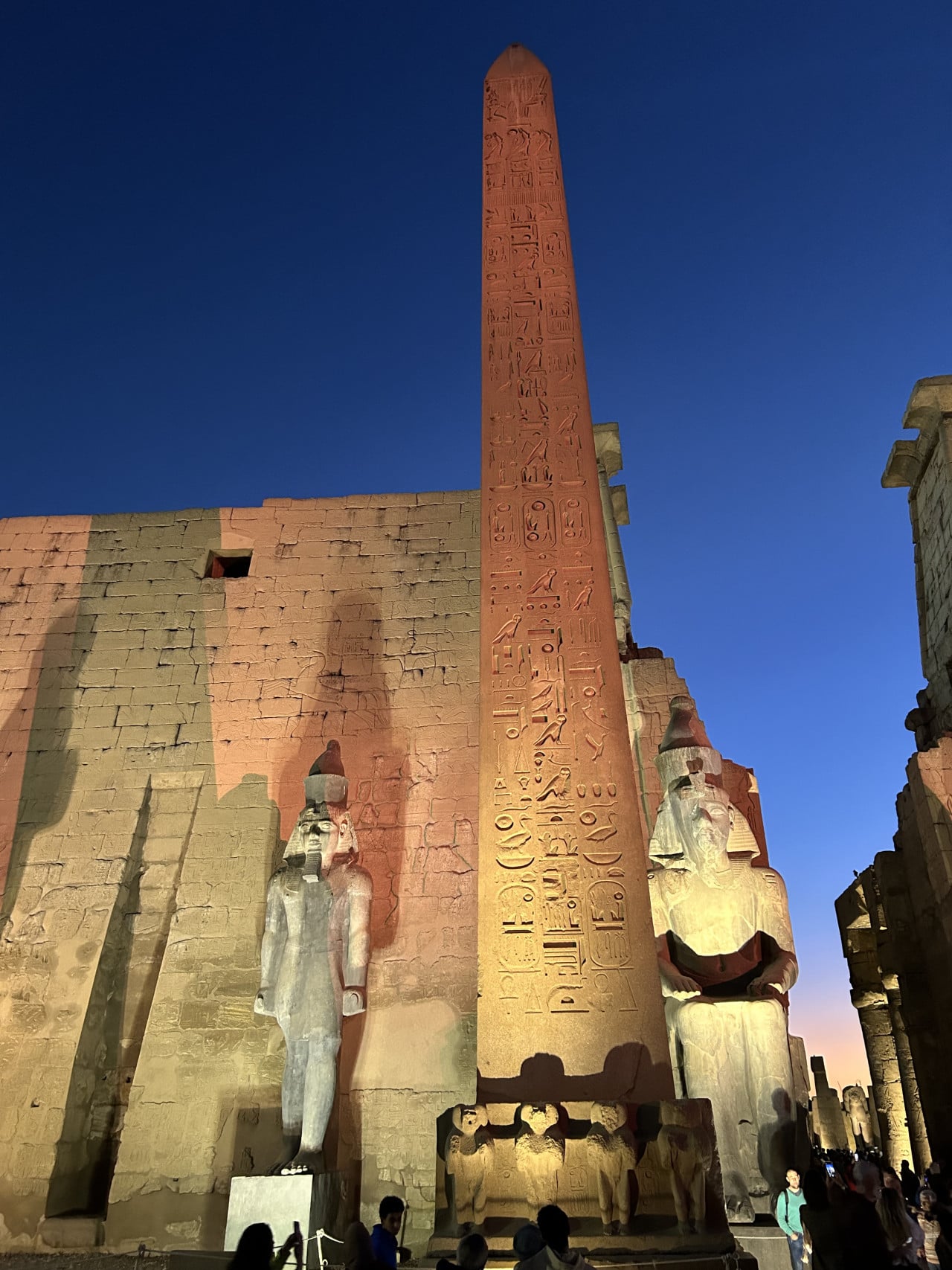 Osvětlený obelisk a dvě obří sochy u chrámové zdi v Karnaku.
