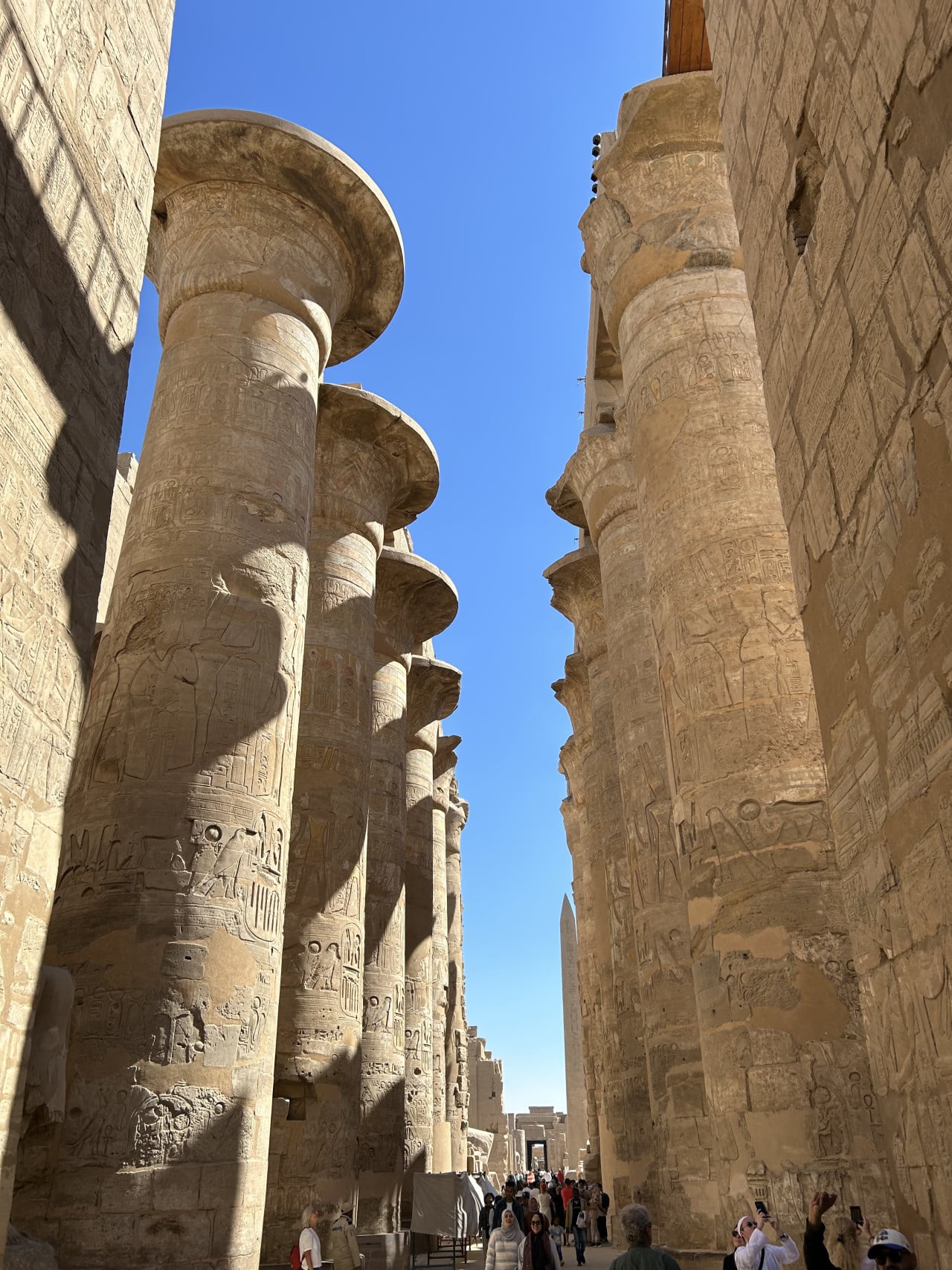Obří sloupy chrámu Karnak s reliéfy a turisty mezi nimi￼