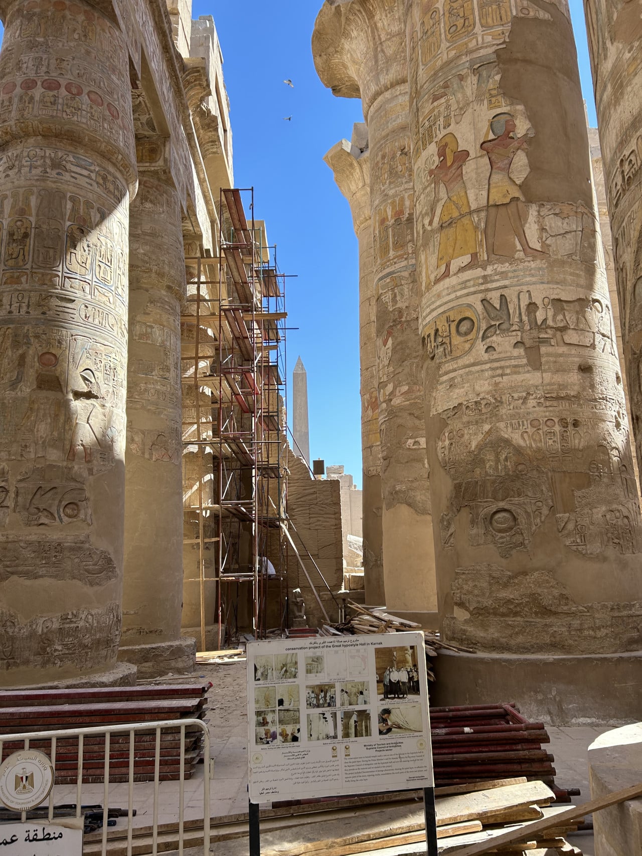 Sloupy chrámu Karnak s hieroglyfy, lešením a informační tabulí pod modrou oblohou