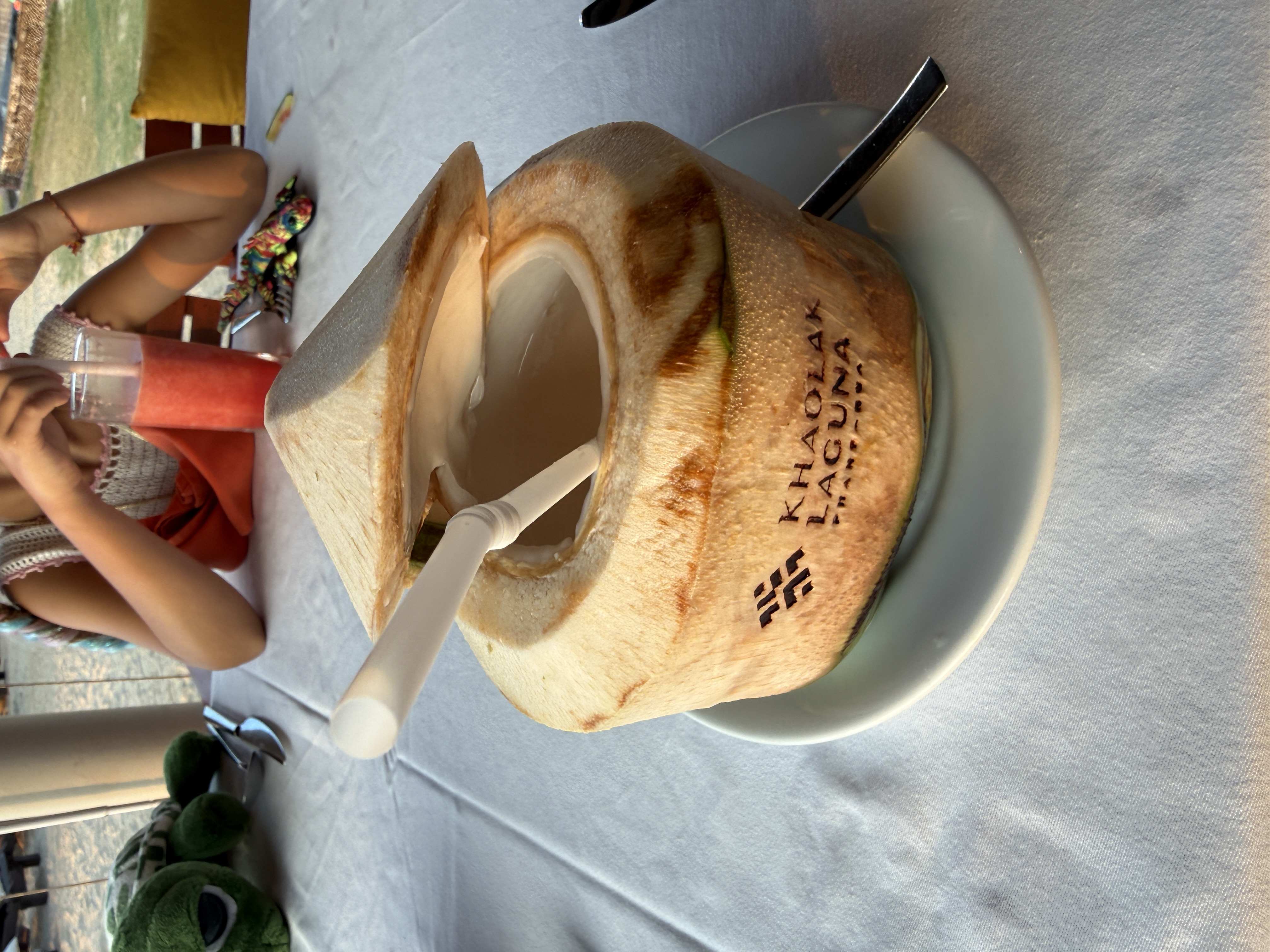 Otevřený kokos s brčkem a logem KHAOLAK LAGUNA RESORT, v pozadí smoothie.