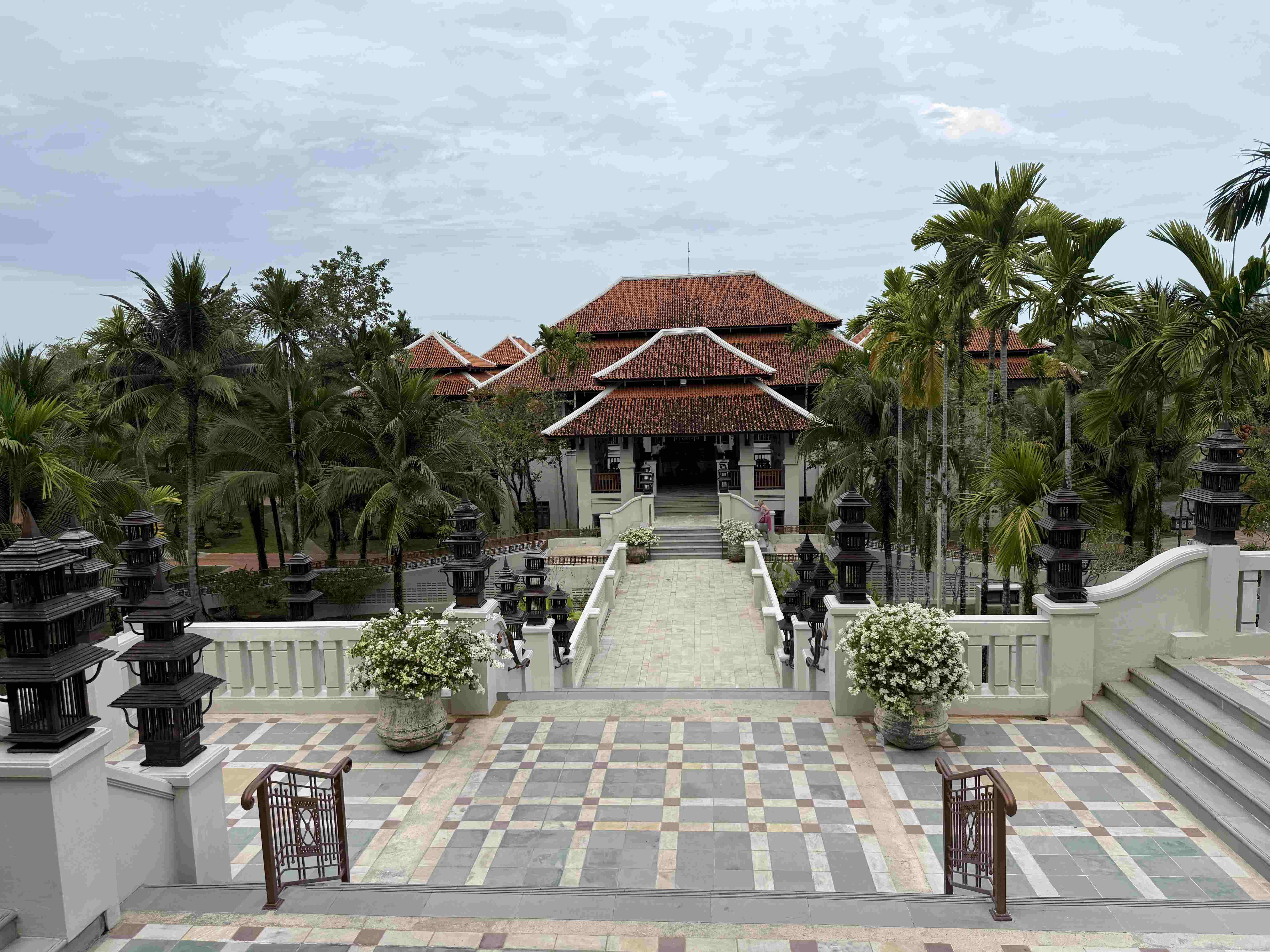 Pohled z terasy na chodník k resortu, červené střechy, palmy a lampy.