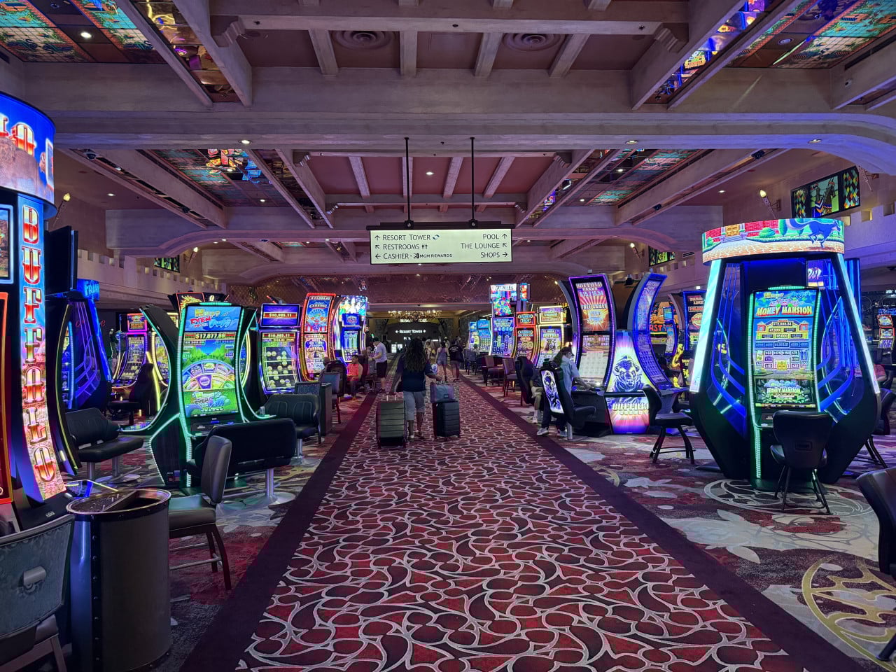 Chodba kasina s automaty, hráči a cedulí Resort Tower, Restrooms, Cashier, Pool, Shops.