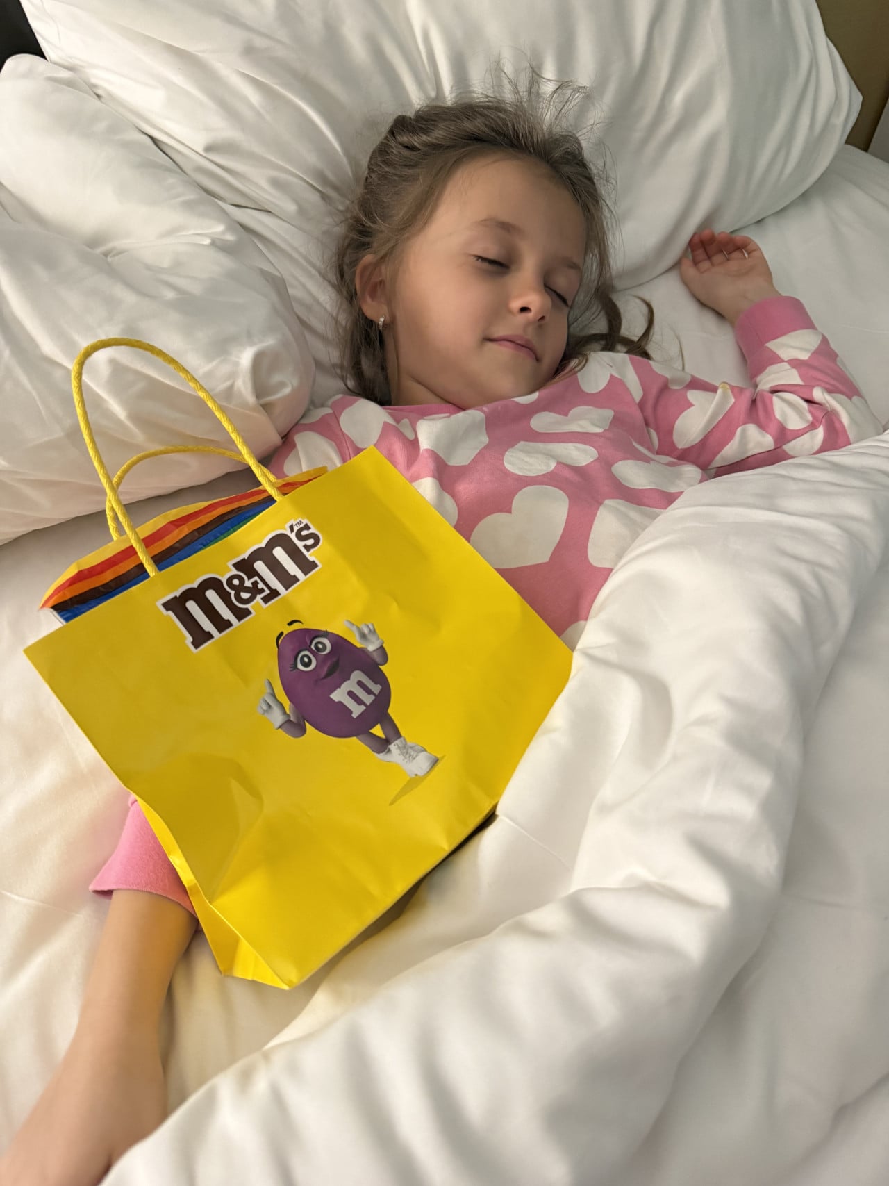 Spící dívka v posteli, na peřině žlutá taška s nápisem m&m’s
