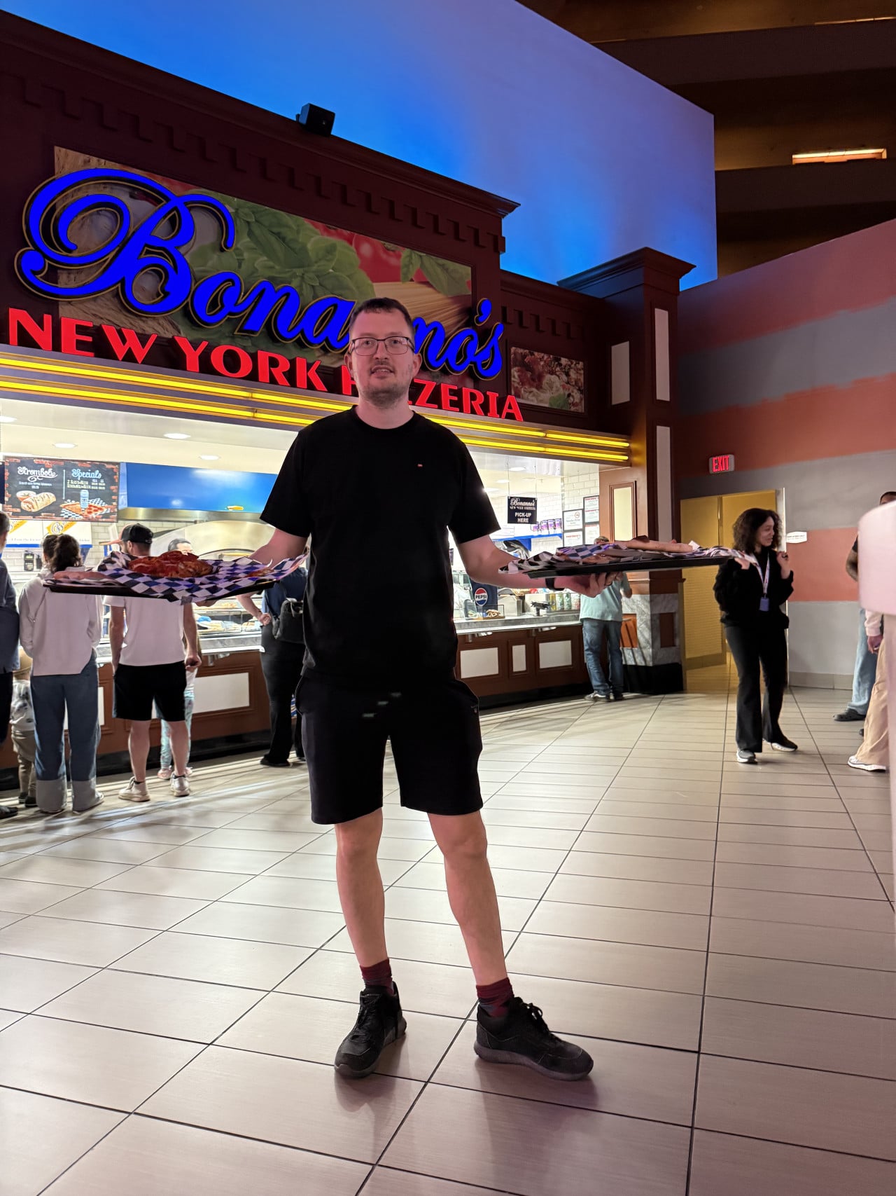 Muž drží dvě tácy s pizzou před nápisem Bonanno's New York Pizzeria