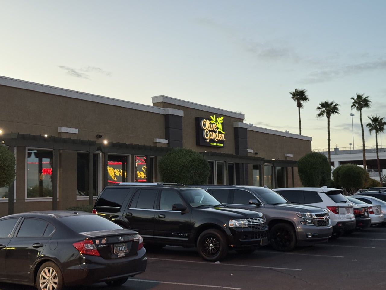 Budova restaurace Olive Garden Italian Kitchen, zaparkovaná auta před ní, palmy za soumraku.