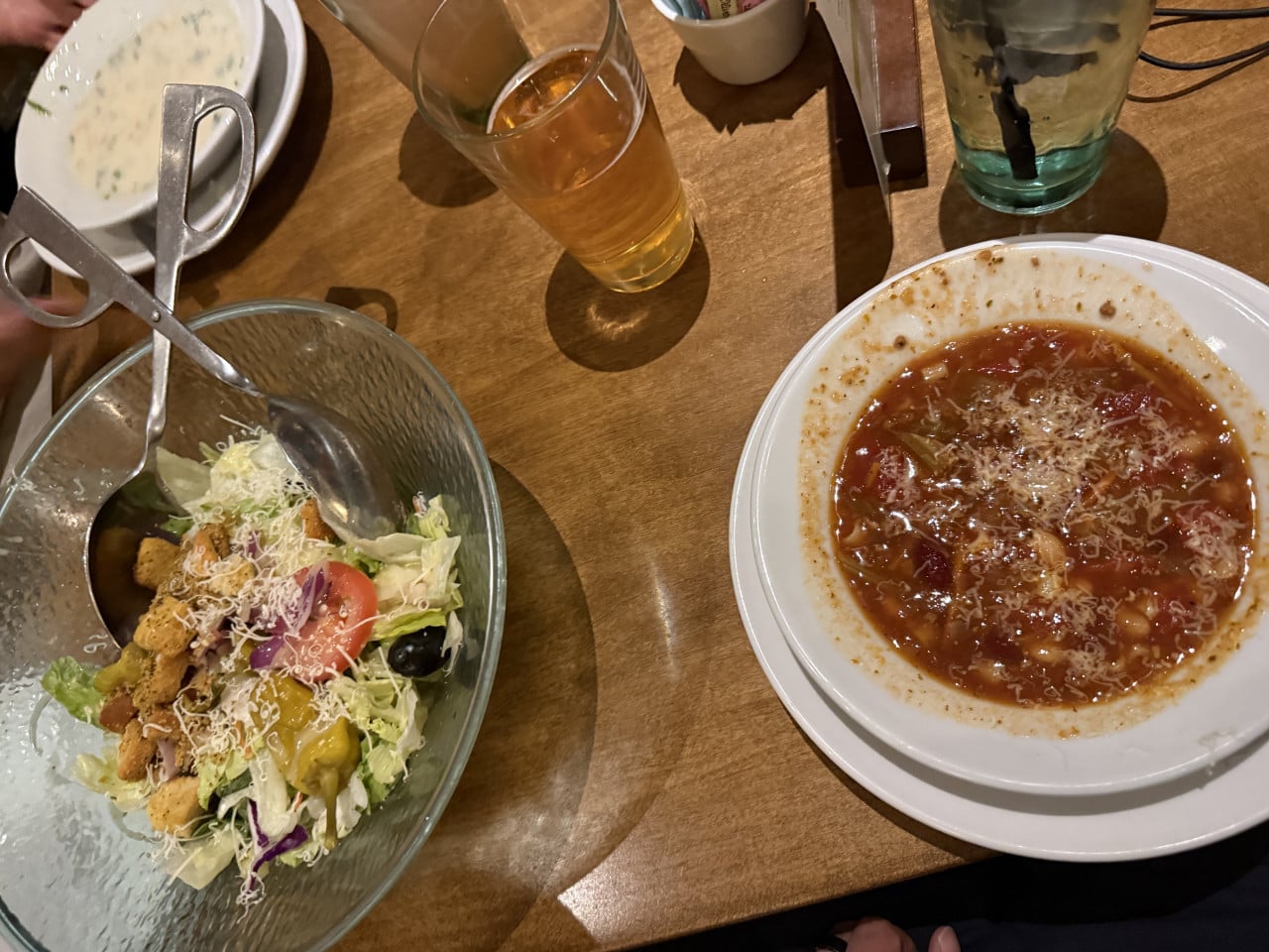 Na dřevěném stole salát v míse, talíř polévky a sklenice piva.