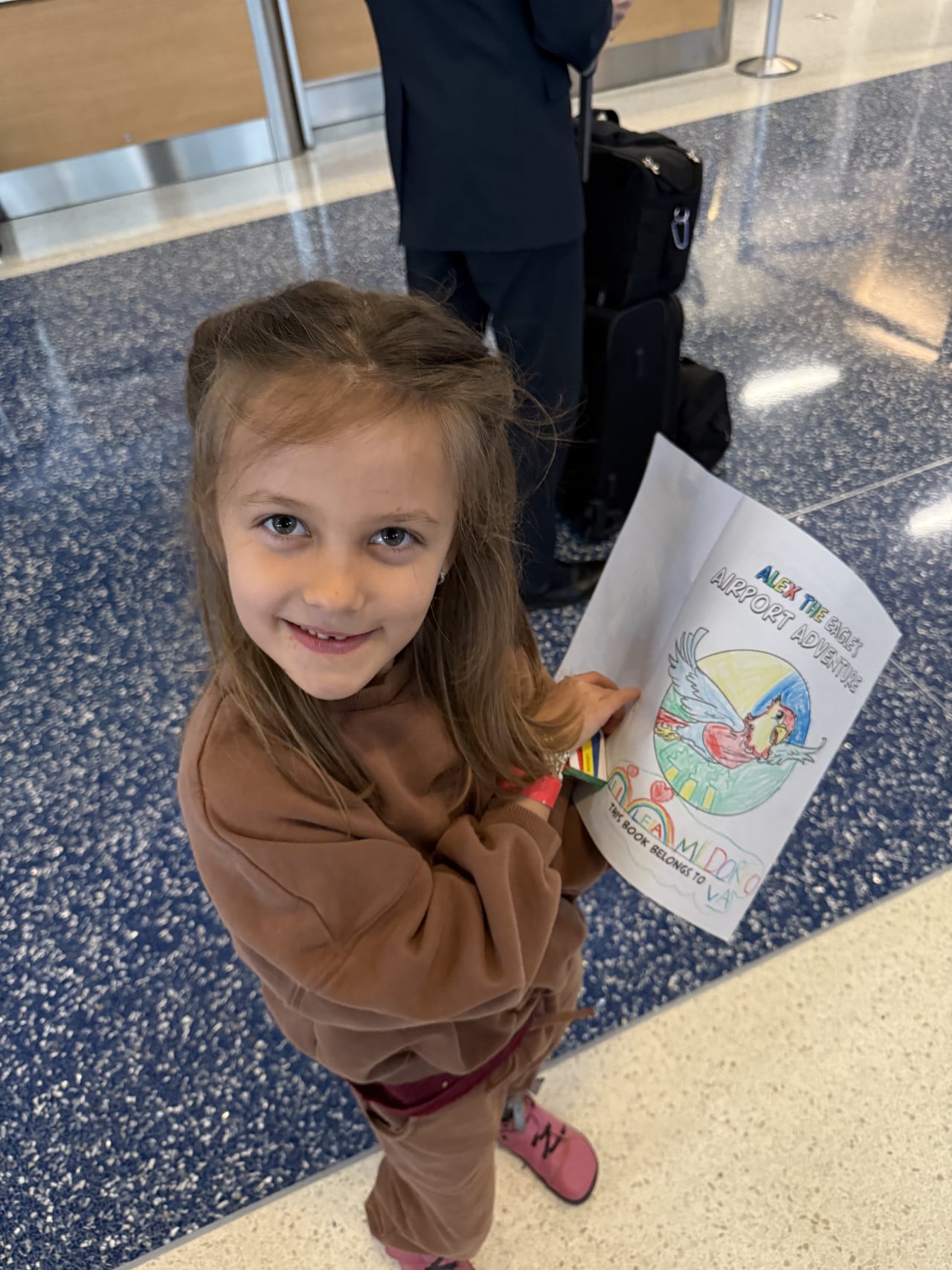 Dívka na letišti drží omalovánku ALEX THE EAGLE'S AIRPORT ADVENTURES, THIS BOOK BELONGS TO