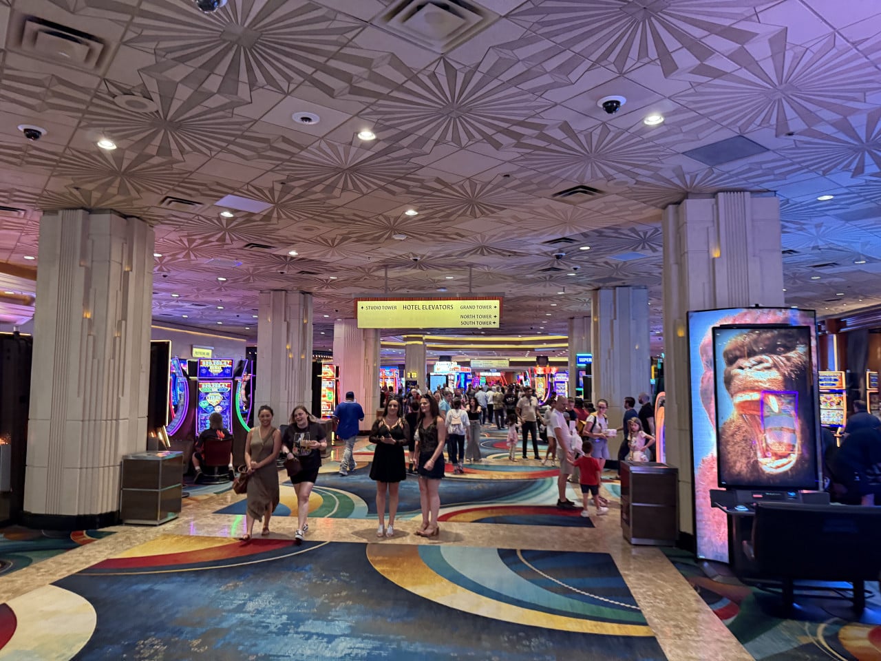 Chodba kasina plná lidí a automatů, cedule HOTEL ELEVATORS uprostřed.