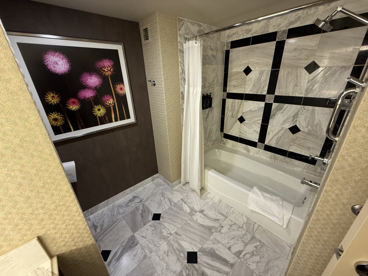 Hotelová koupelna s mramorovou podlahou, vanou, sprchovým závěsem a obrazem květin.