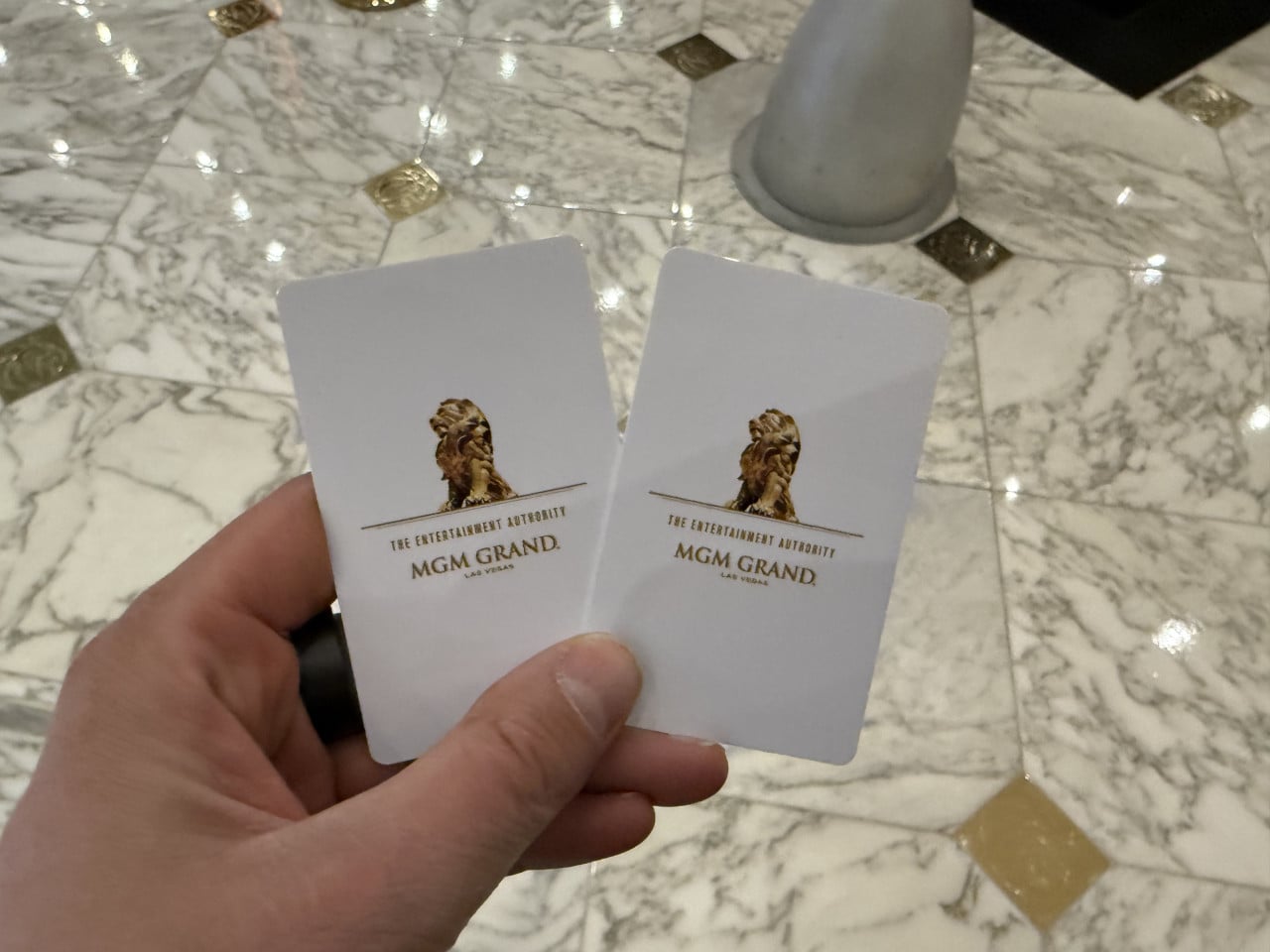 Ruka drží dvě hotelové karty The Entertainment Authority, MGM GRAND Las Vegas, na mramorové podlaze.