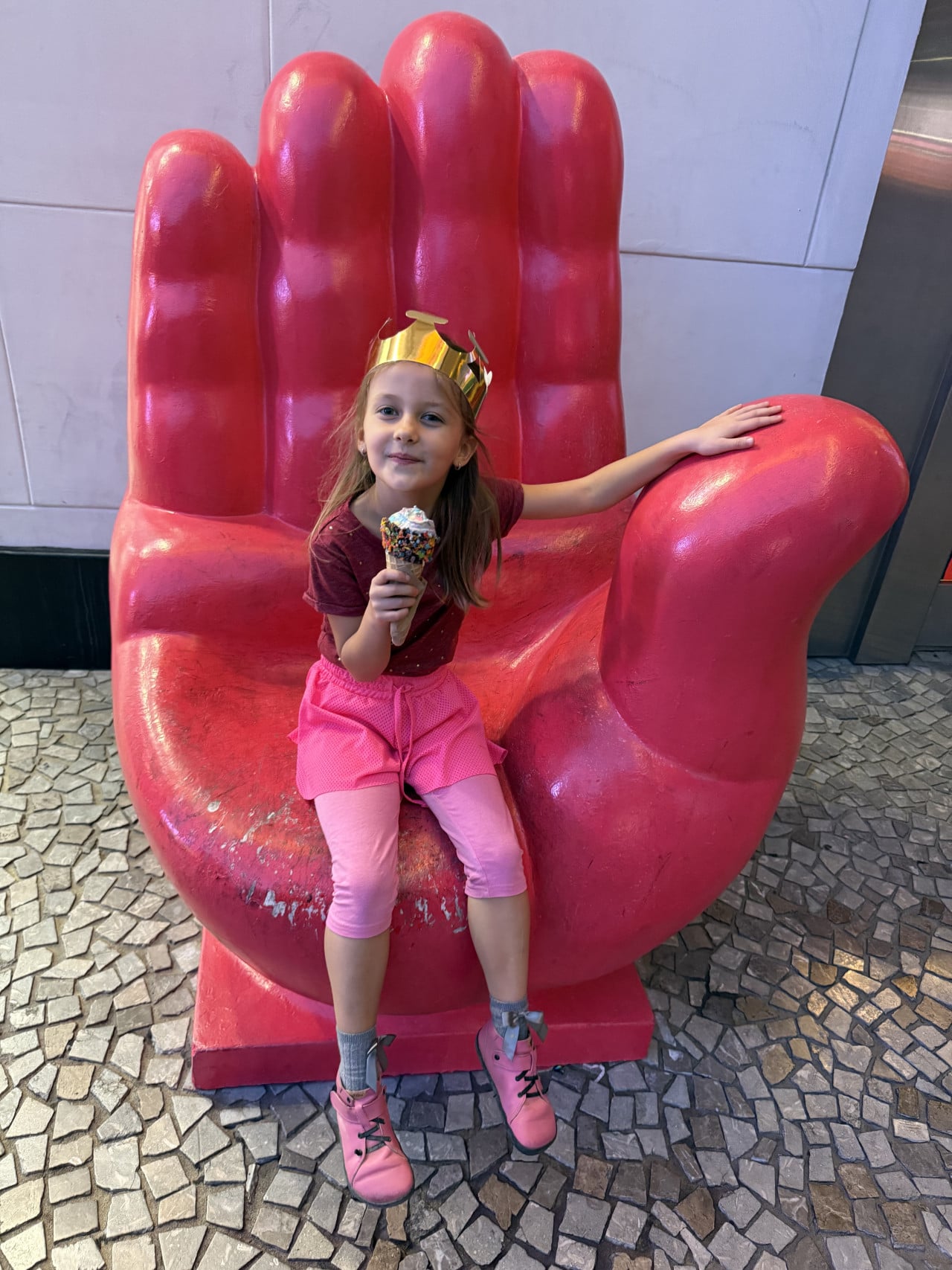 Dívka s papírovou korunou jí zmrzlinu na obřím červeném křesle ve tvaru ruky.