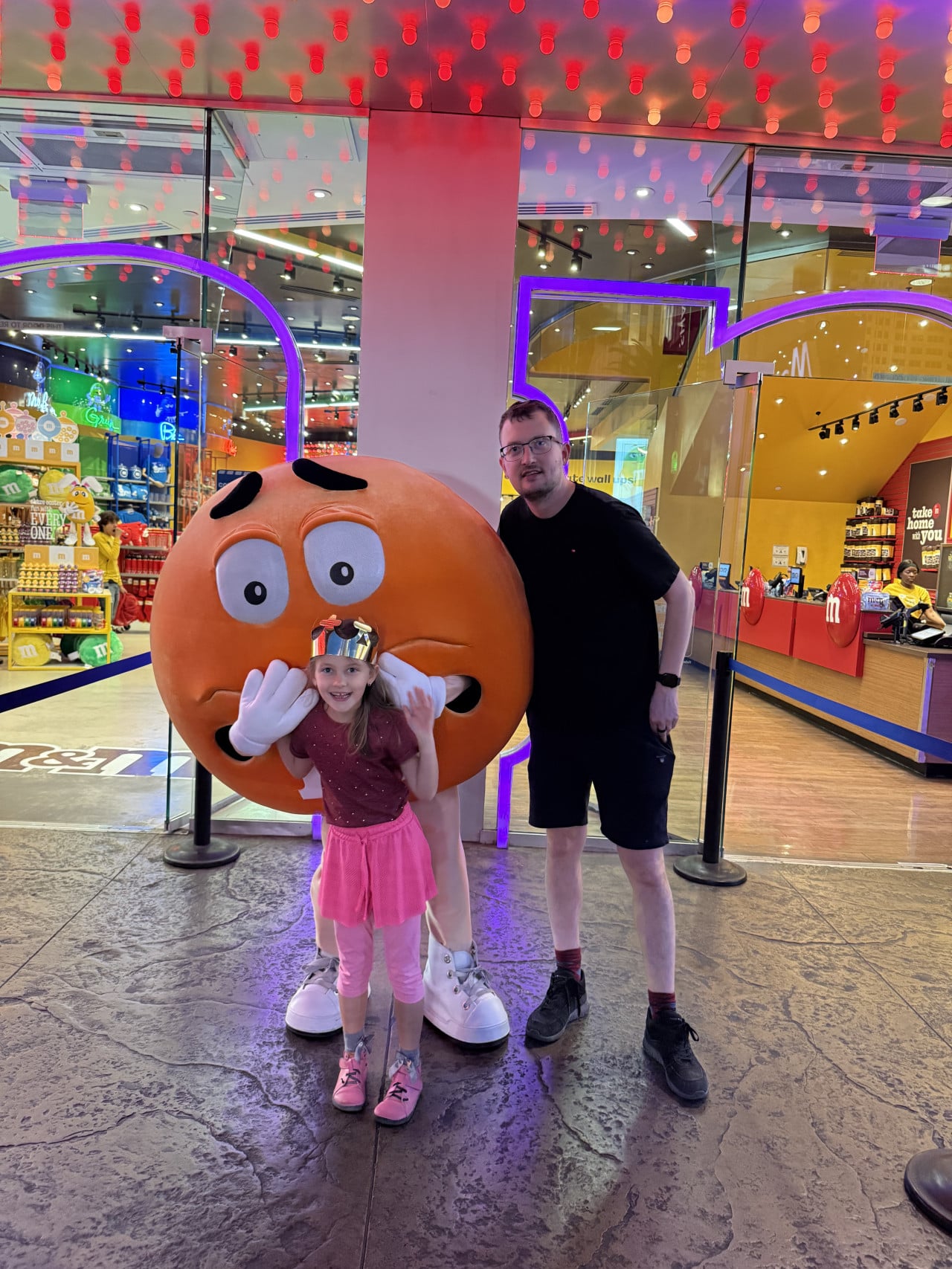 Dívka a muž pózují s oranžovým M&M, v pozadí nápisy m&m's