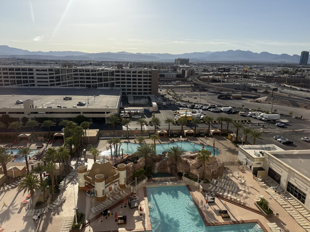 Výhled na hotelový bazén s palmami, lehátky, parkovištěm a horami v dálce