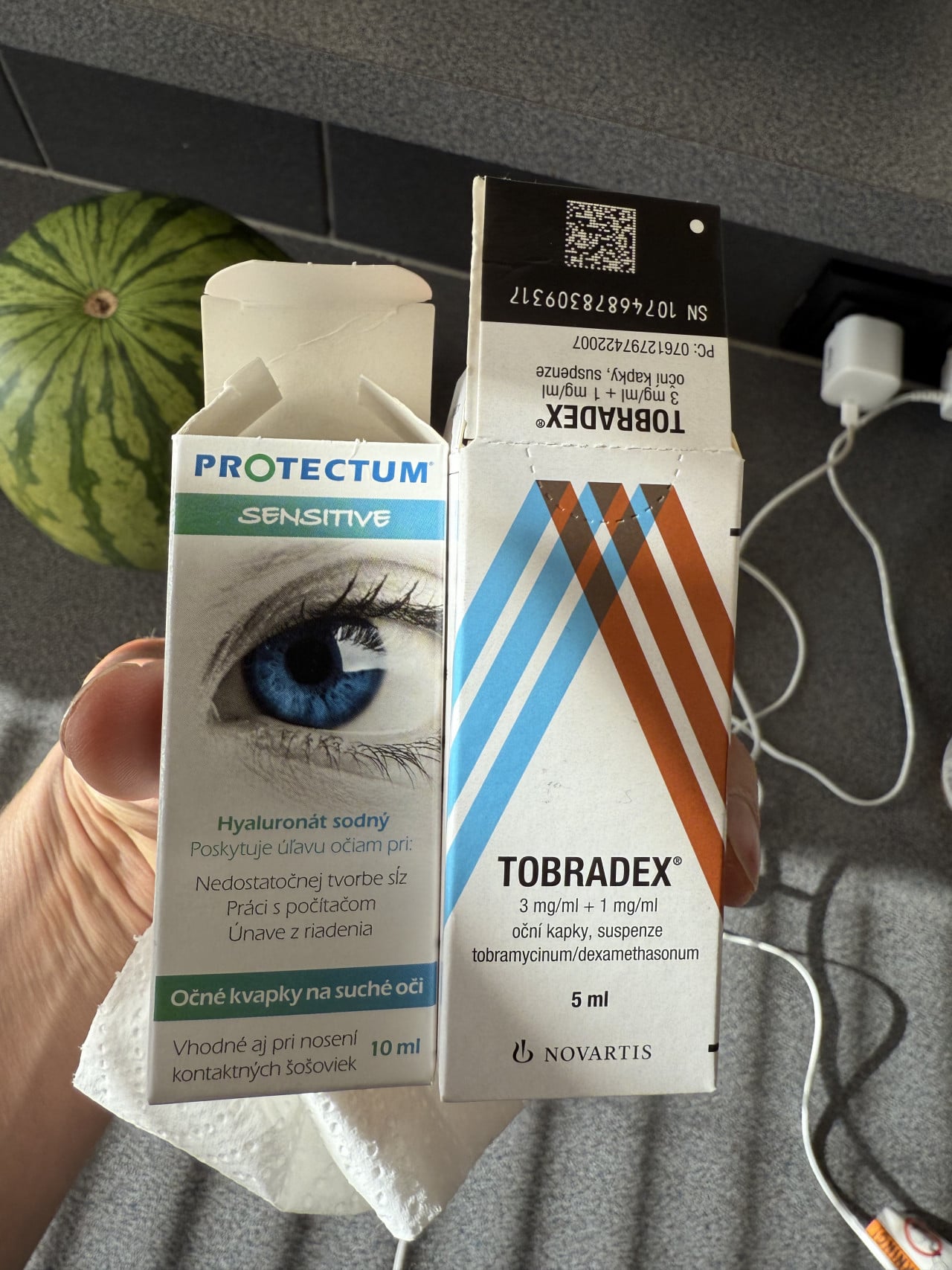 Oční kapky ProtecTum Sensitive a Tobradex v ruce, na pozadí meloun a kabely.